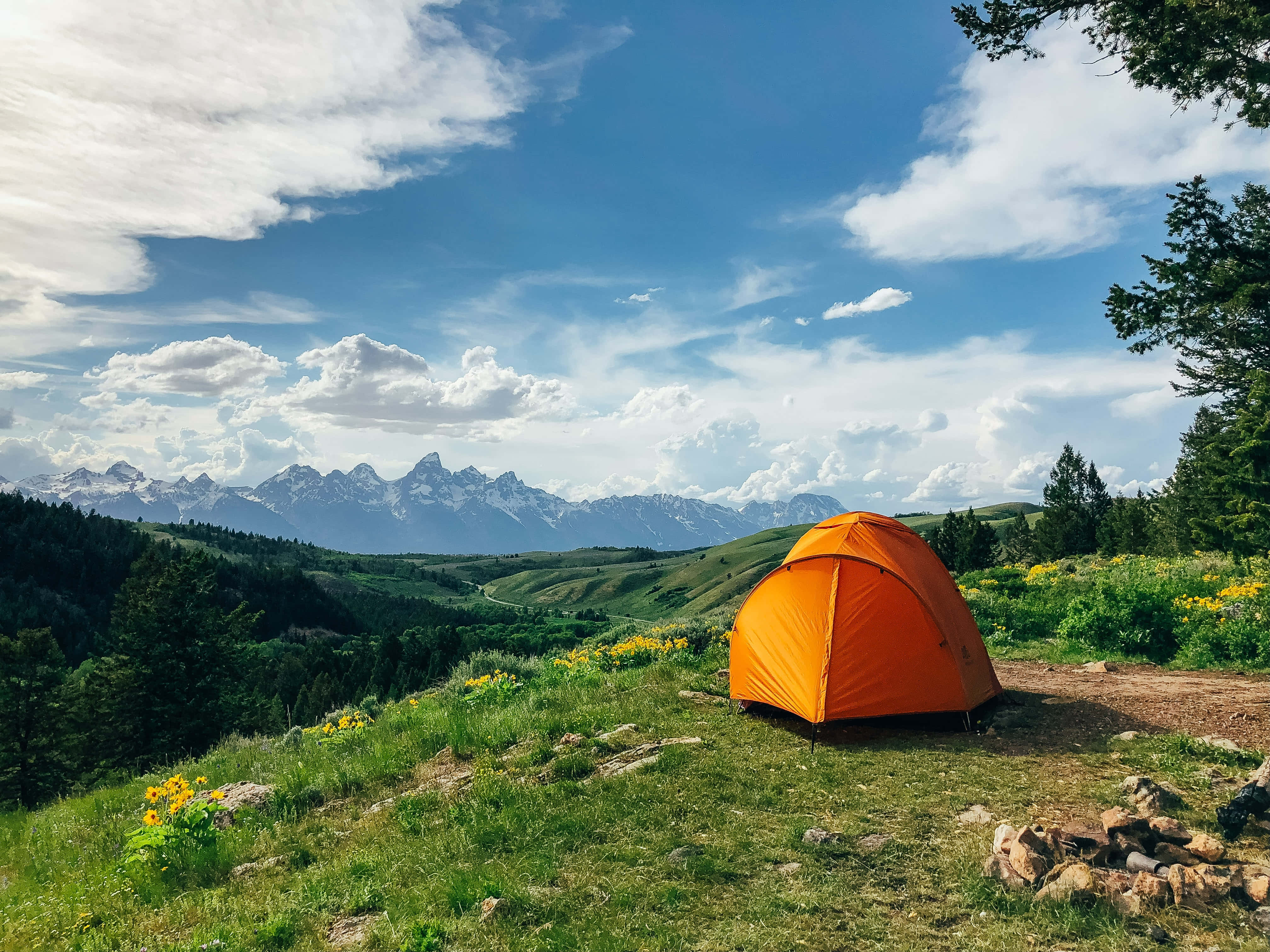 Camping 桌布 壁紙 — 一頂亮橙色的帳篷獨自矗立在青草山坡上，俯瞰著部分多雲的藍天下雄偉的雪山山脈。