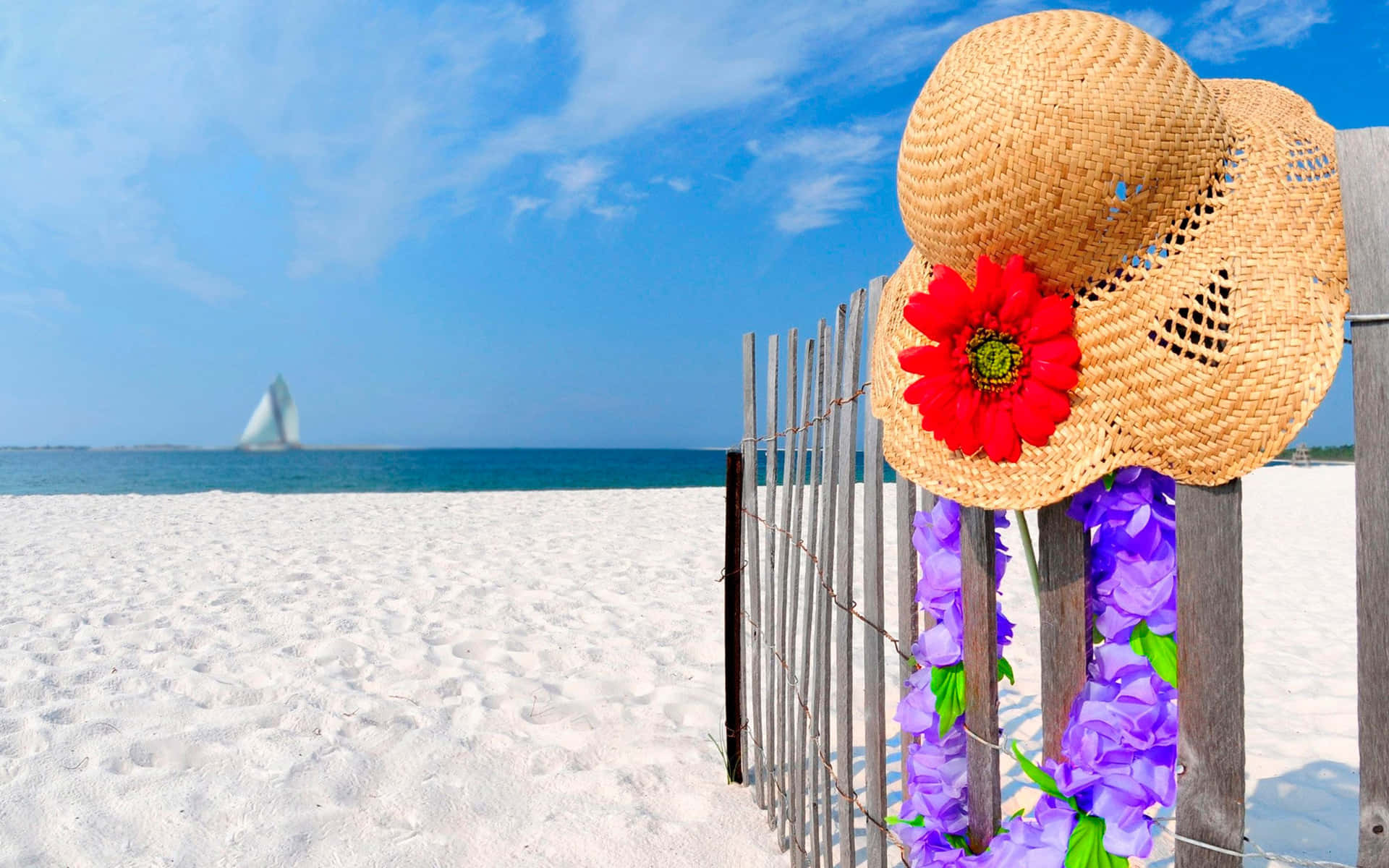 Beach Hat 桌布 壁紙 — 一頂帶有紅花和紫色花環的草帽掛在木柵欄上，俯瞰著白色沙灘，遠處有一艘帆船，在晴朗的藍天下。