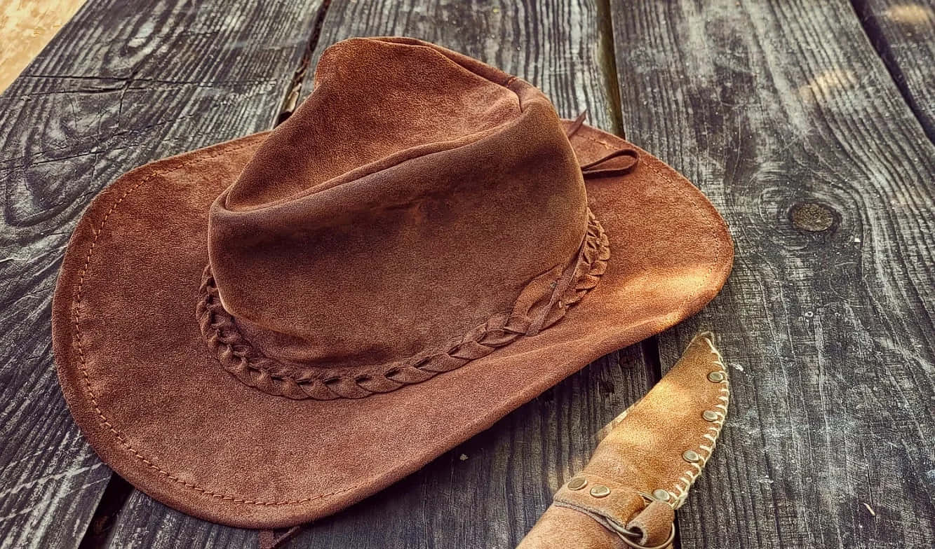 Cowboy Hat 桌布 壁紙 — 一頂棕色麂皮牛仔帽，配有編織皮革帶，旁邊放著一把皮革鞘刀，擱在風化的木板上。