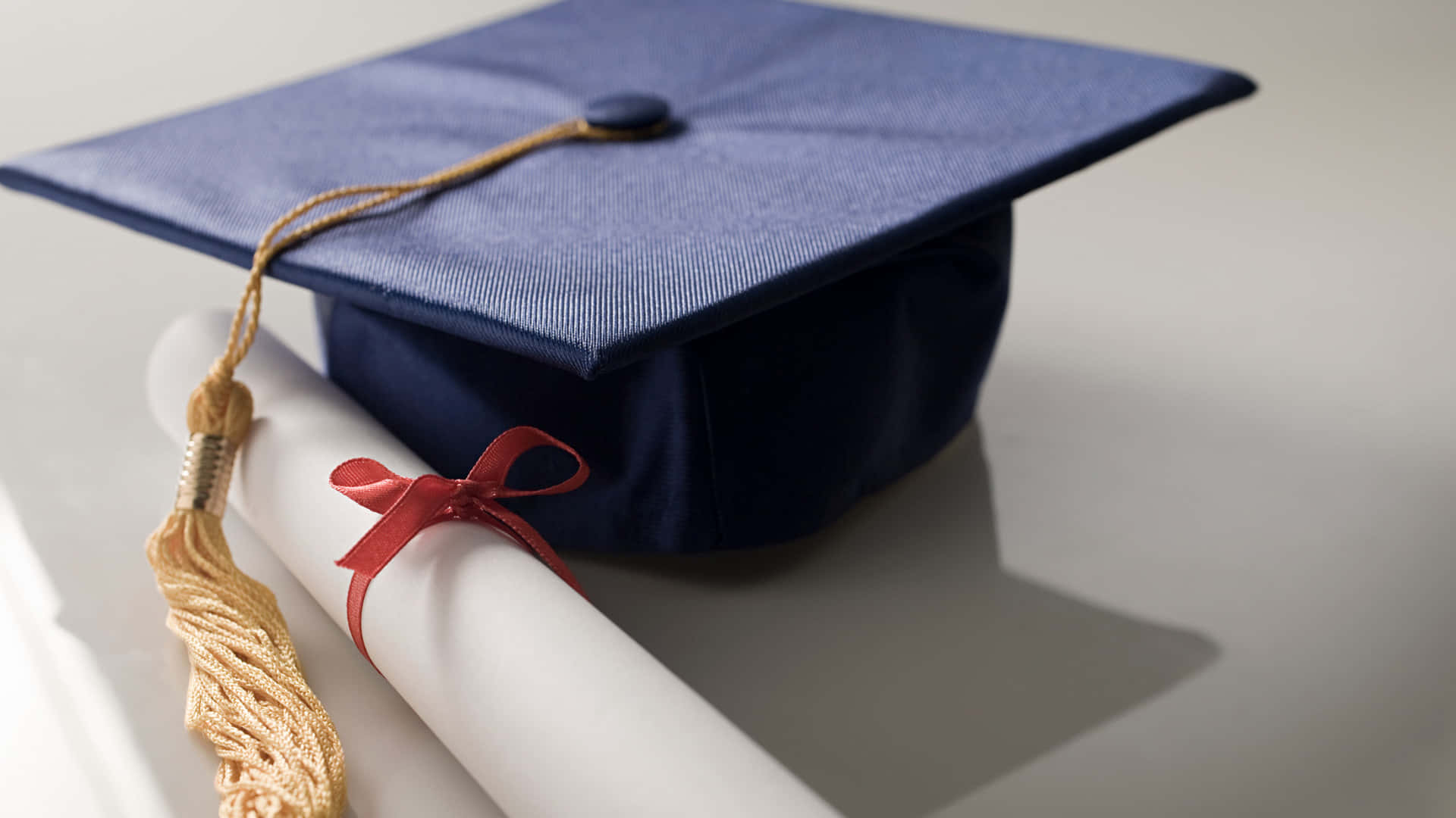Graduation Ceremony 桌布 壁紙 — 一頂海軍藍色畢業帽搭配金色流蘇，旁邊放著一份用紅絲帶綁著的白色捲起畢業證書，置於潔白表面上。