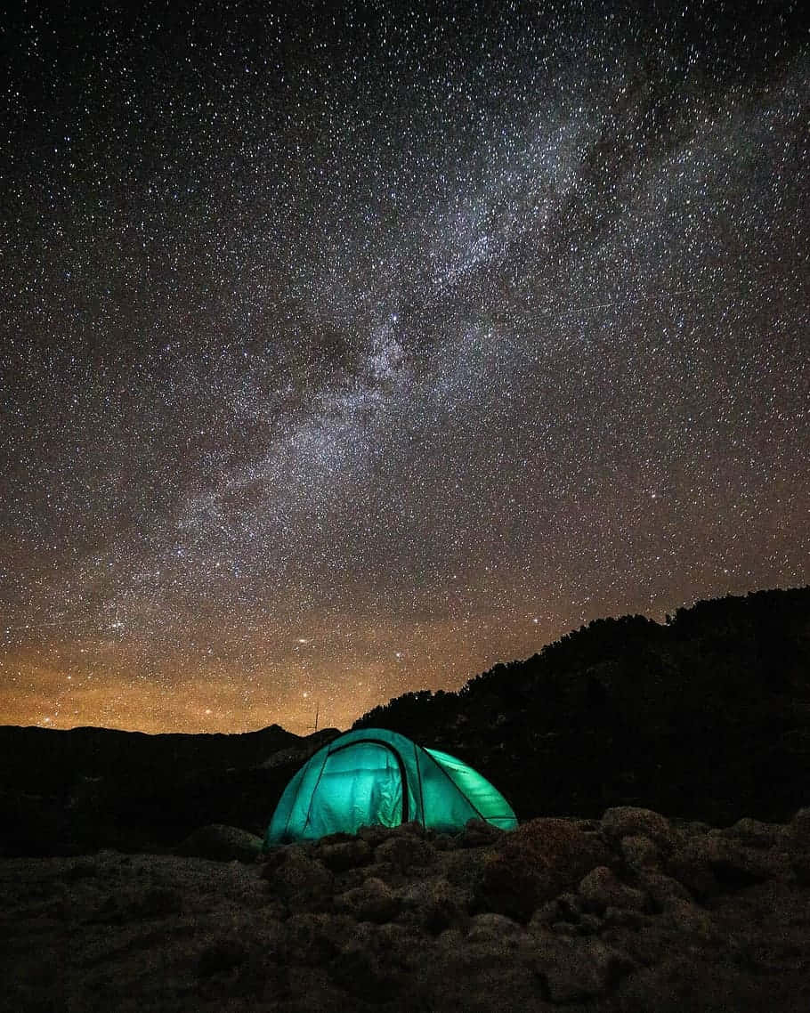Light 桌布 壁紙 — 一頂發光的綠色帳篷 nestled 在岩石之間，上方是令人嘆為觀止的星空，銀河拱跨天際。