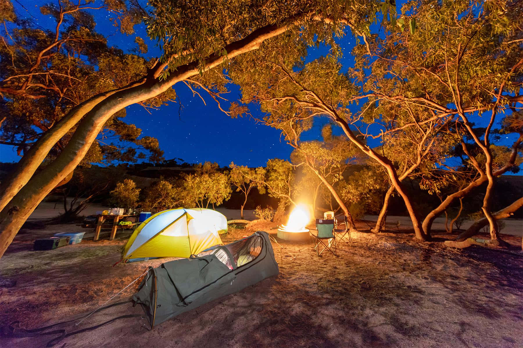 Camping 桌布 壁紙 — 一頂發光的黃色帳篷和睡袋在繁星點點的夜空下，旁邊是劈啪作響的營火，周圍環繞著蔓延的樹木。