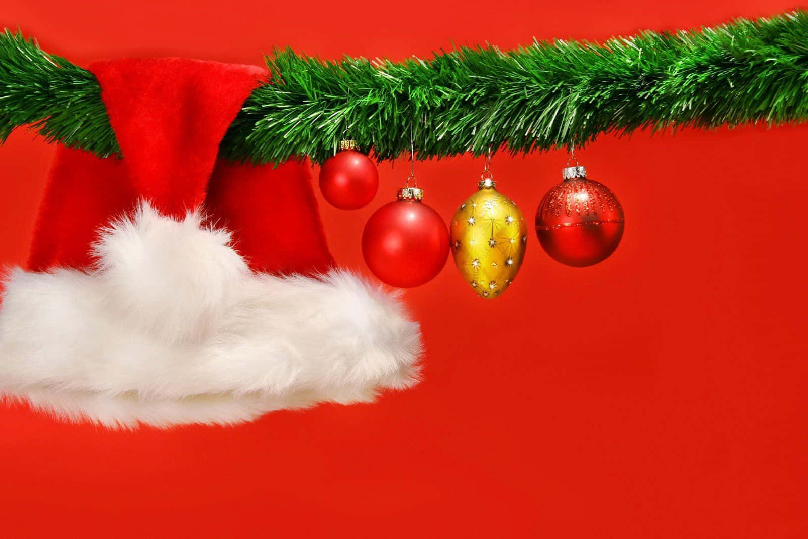 Santa Claus 桌布 壁紙 — 一頂紅色聖誕帽，帶有白色毛邊，放在一個掛有紅色和金色裝飾品的綠色花環下，背景是鮮豔的紅色。