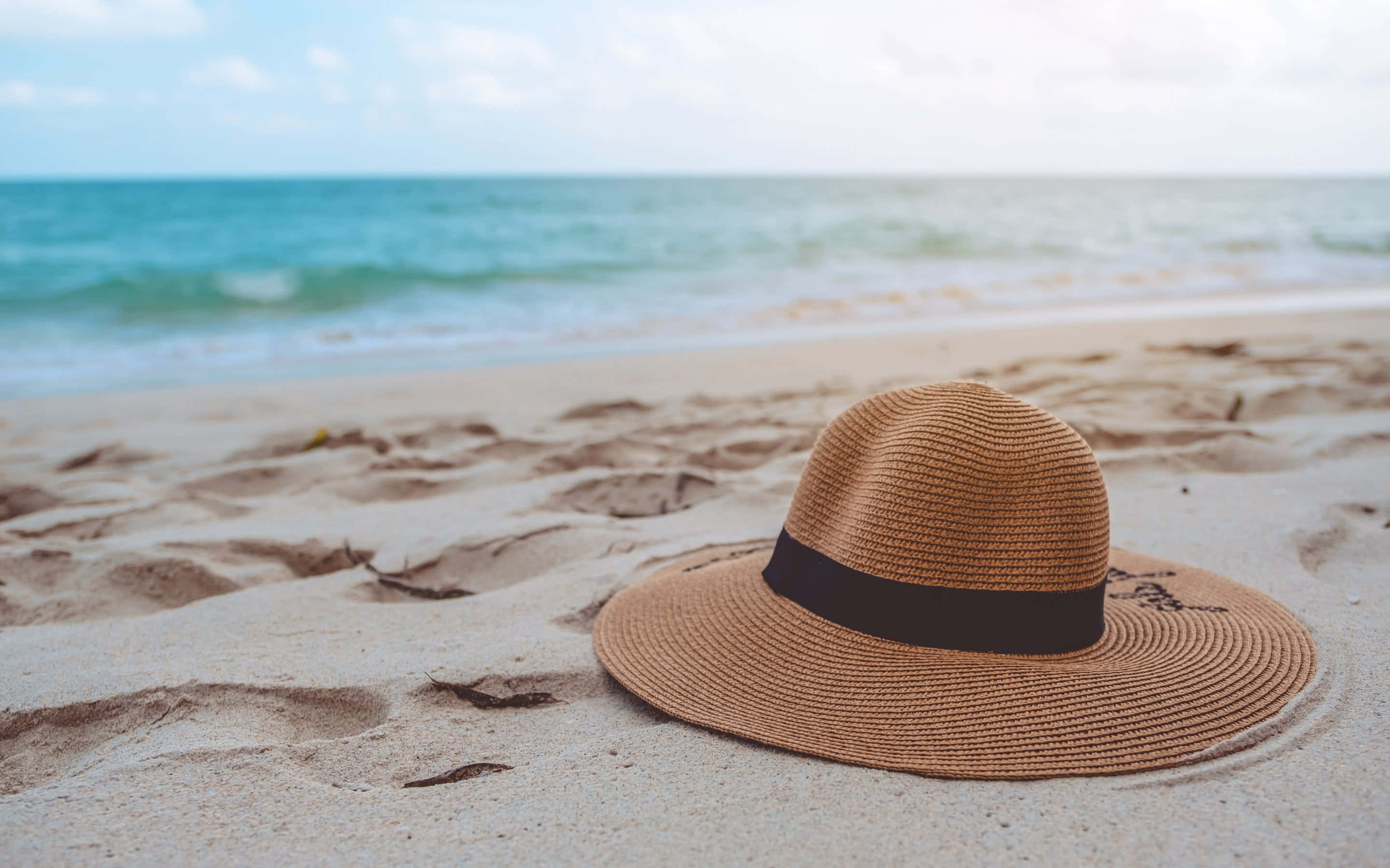 Beach Hat 桌布 壁紙 — 一頂編織草帽，帶有黑色帶子，擱在質感沙灘上，旁邊是碧綠的海洋，天空明亮。