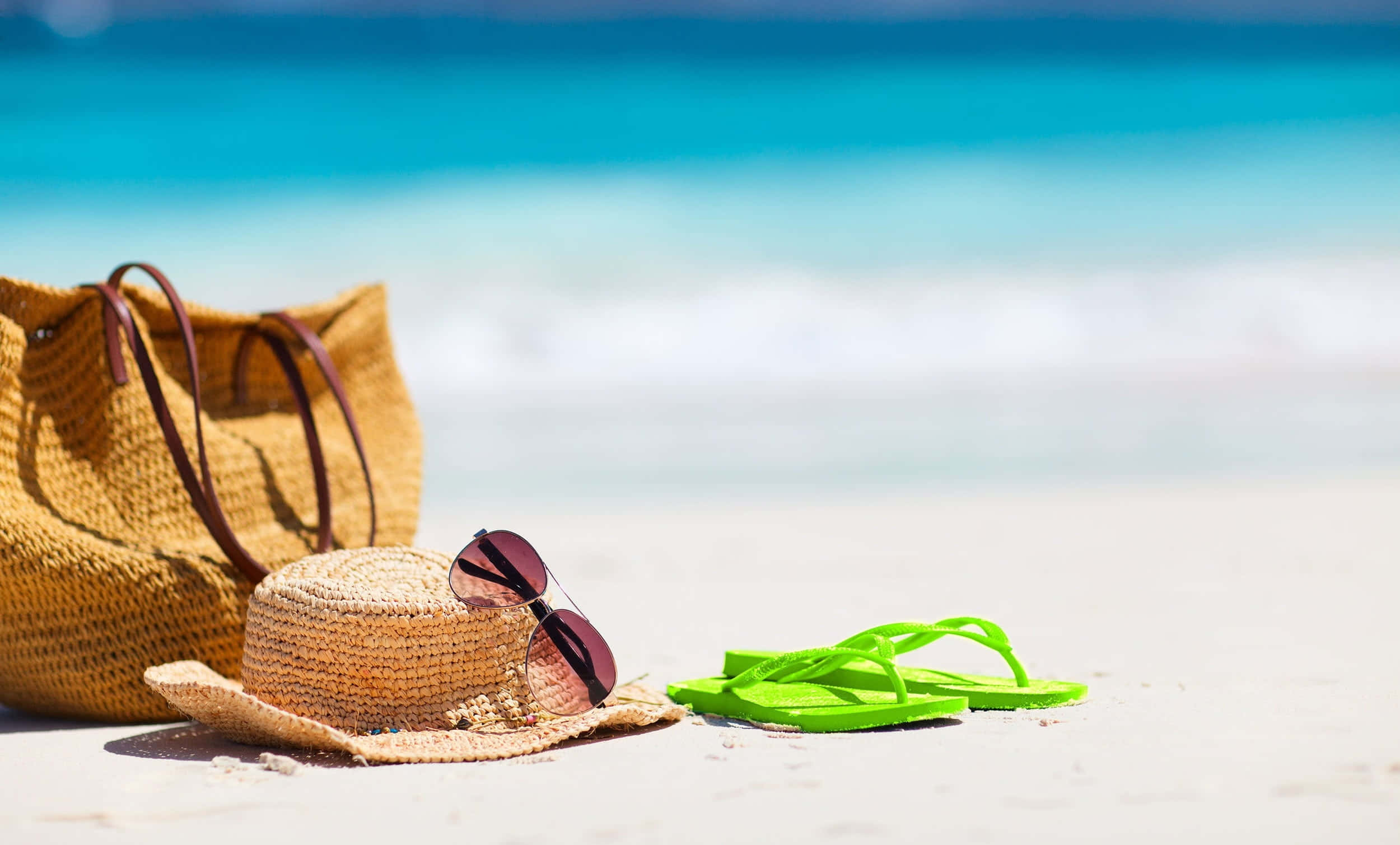 Beach Bag 桌布 壁紙 — 一頂草帽、太陽眼鏡和亮綠色夾腳拖放在白色沙灘上，旁邊有一個編織袋，背景是藍綠色的海浪。