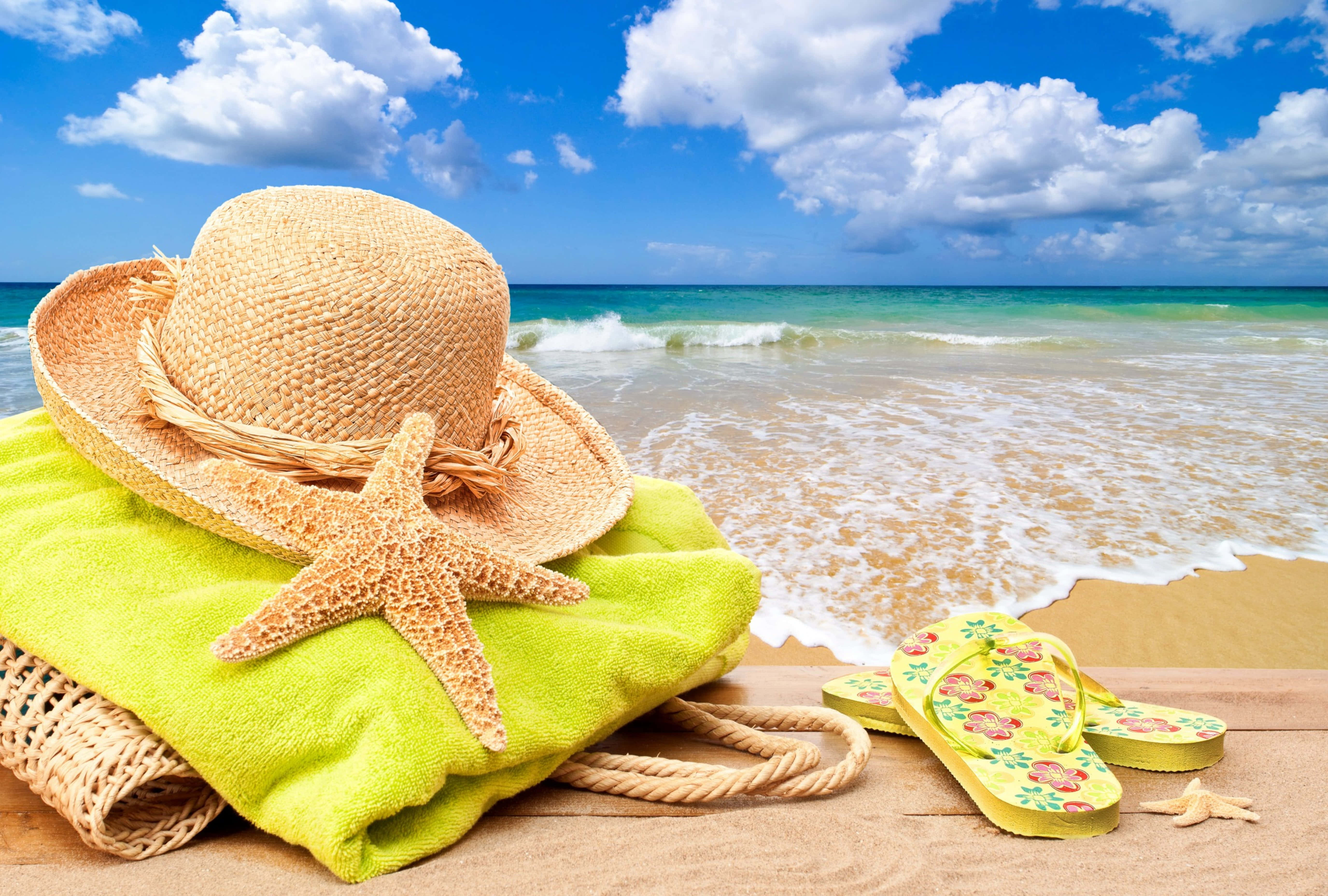Beach Accessories 桌布 壁紙 — 一頂草帽、海星和花卉夾腳拖鞋放在亮綠色毛巾上，旁邊是海浪，天空蔚藍，白雲朵朵。