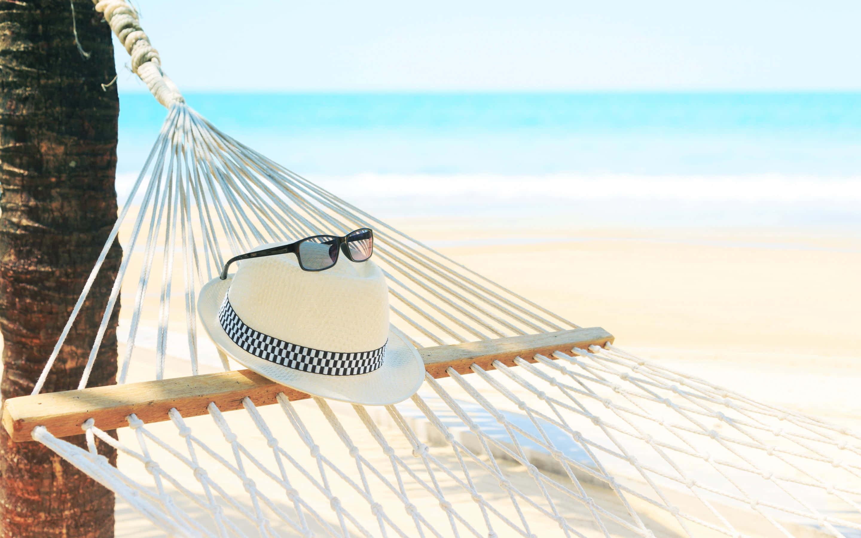 Beach Hat 桌布 壁紙 — 一頂草帽和太陽眼鏡放在一張白色吊床上，吊床繫在棕櫚樹之間，俯瞰著碧綠的海洋和沙灘。