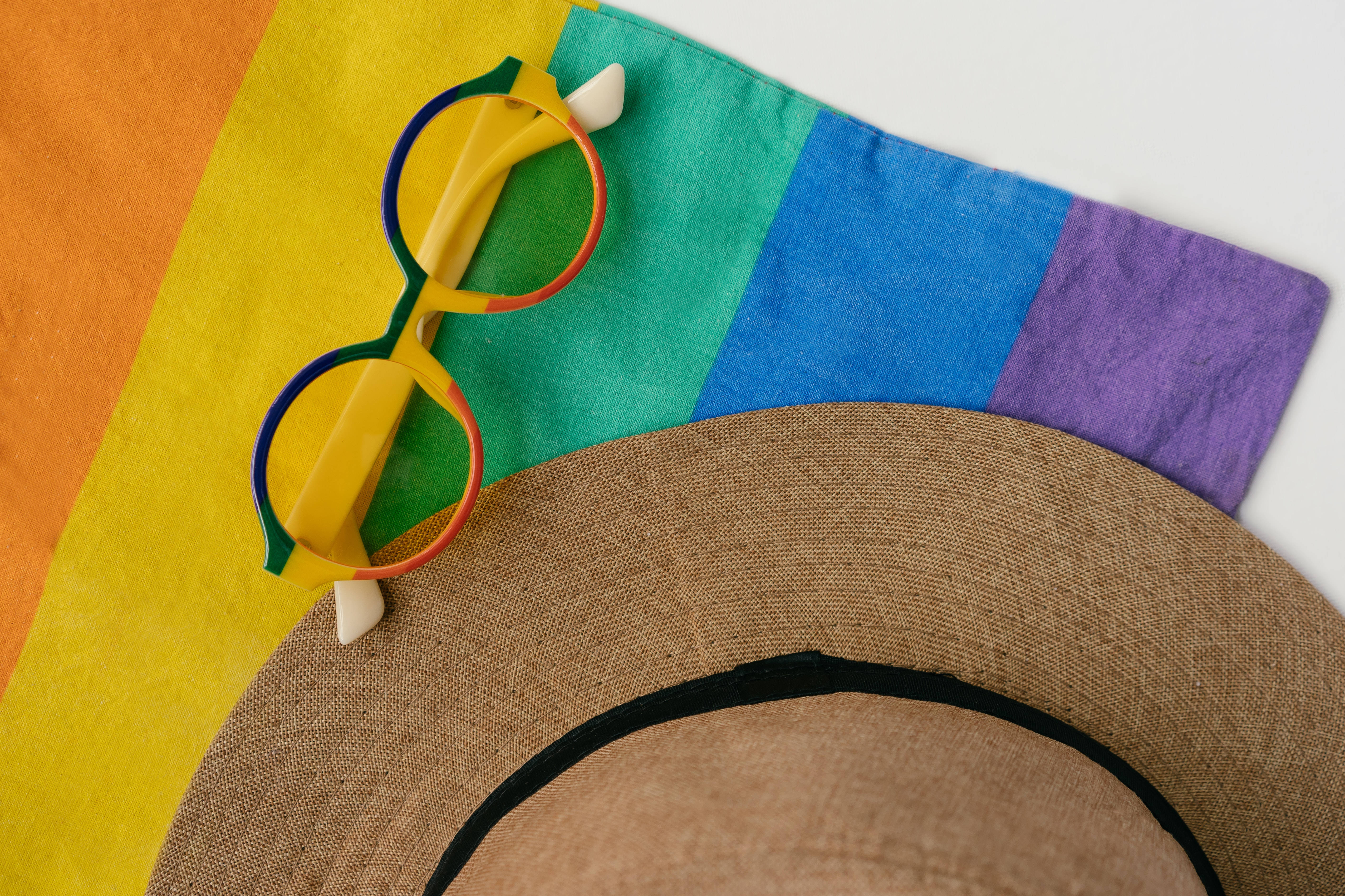 Pride Flag 桌布 壁紙 — 一頂草帽和彩色彩虹眼鏡放在鮮豔的彩虹旗布料上。