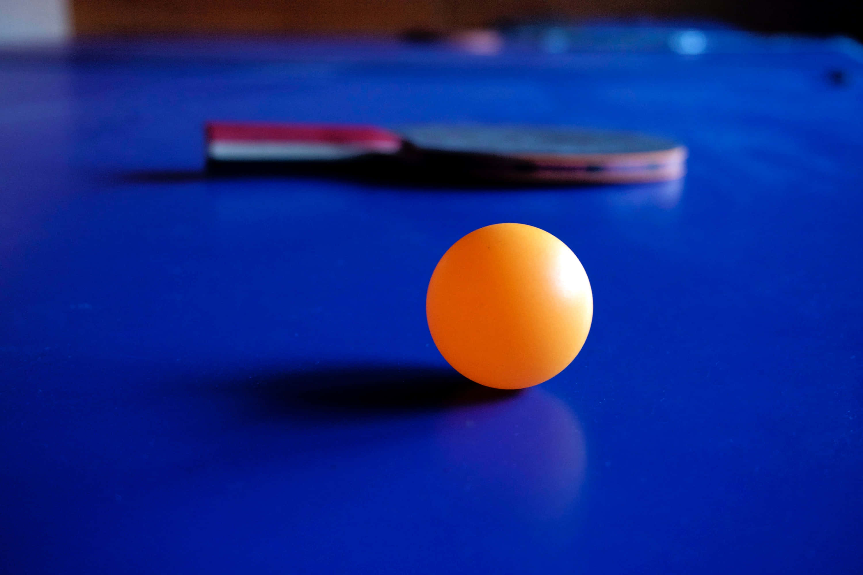 ball 桌布 壁紙 — 一顆亮橙色的乒乓球清晰聚焦在深藍色的桌子上，背景有一塊模糊的球拍。