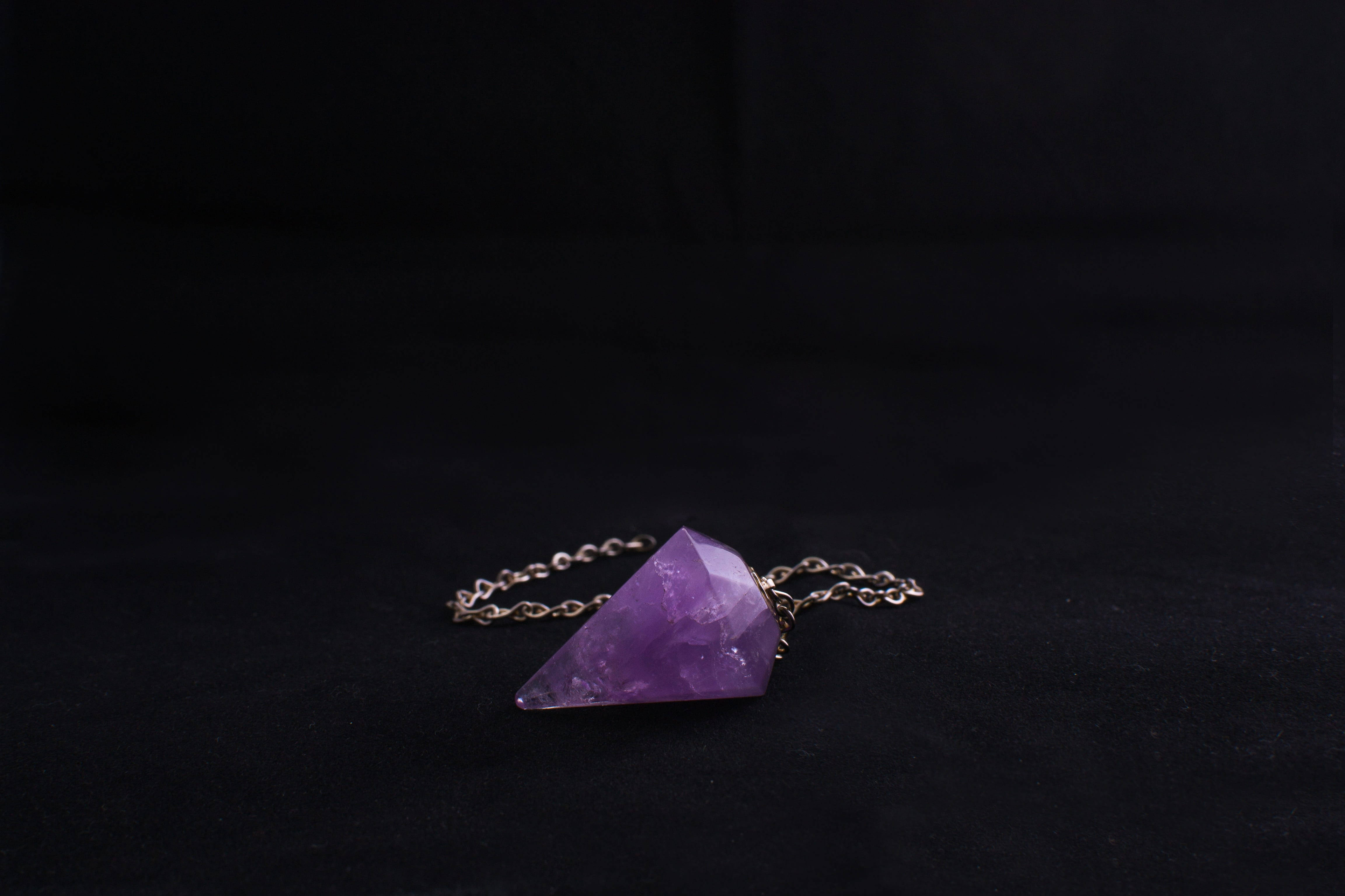 紫水晶 桌布 壁紙 — 一顆刻面紫色紫水晶擺錘放在深色天鵝絨表面上，金屬鏈條纏繞在旁邊。