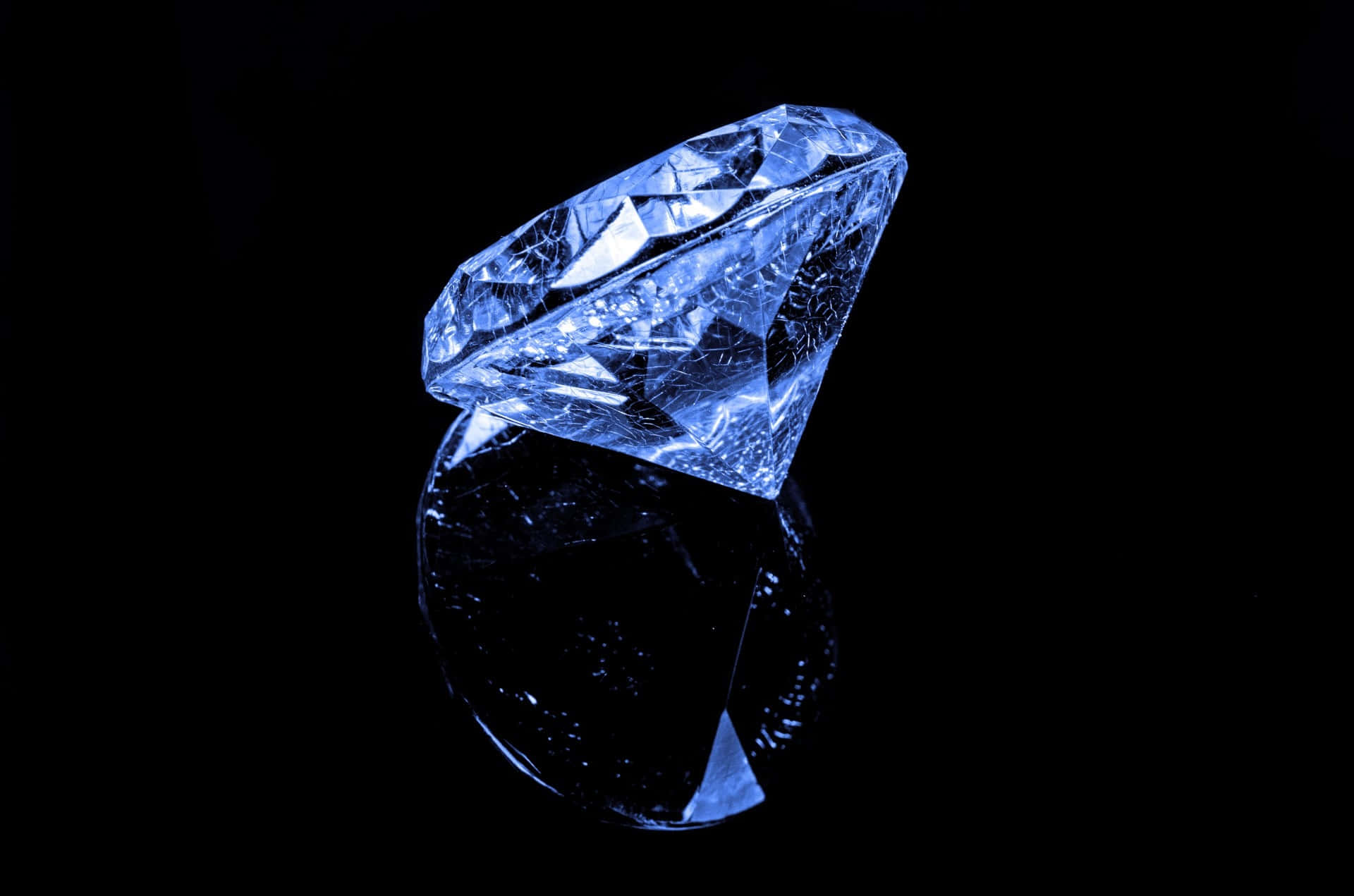Blue Diamonds 桌布 壁紙 — 一顆單一的藍色調鑽石在黑色反光表面上閃耀著銳利的切面，投射出鏡面般的光澤。