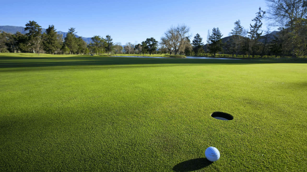 Golf Ball 桌布 壁紙 — 一顆完美無瑕的高爾夫球停在距離球洞僅幾英吋的位置，球道陽光普照、綠意盎然，遠方有樹木和山脈環繞。