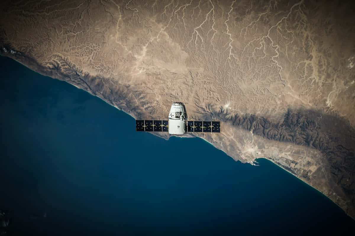 Telescope 桌布 壁紙 — 一顆帶有太陽能板的衛星在崎嶇的海岸線上空運行，乾旱的陸地與深藍色的海洋在此交匯。