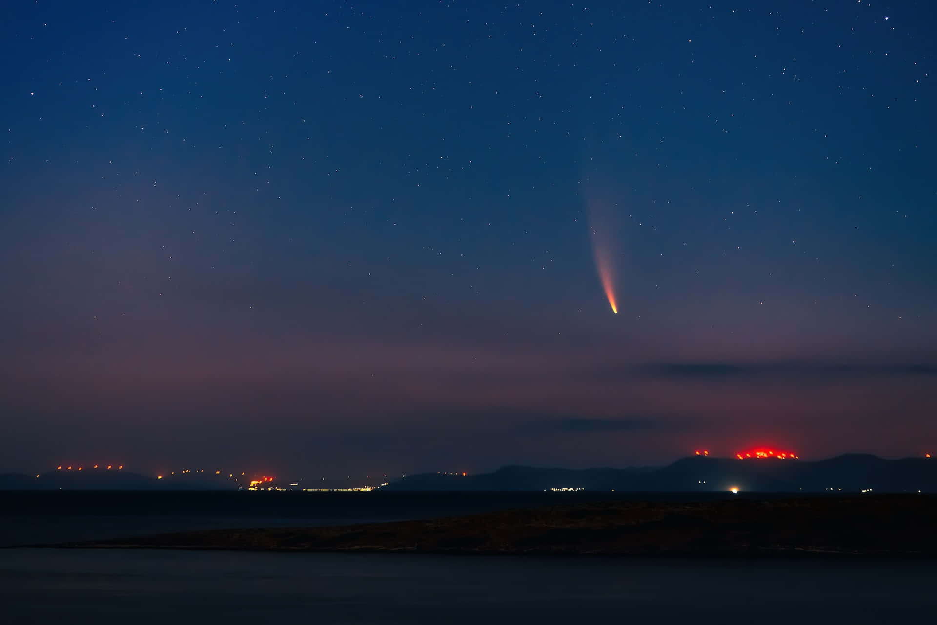 comet 桌布 壁紙 — 一顆彗星劃過繁星點點的暮色天空，下方是黑暗的沿海景觀，遠處有紅色和白色的燈光。