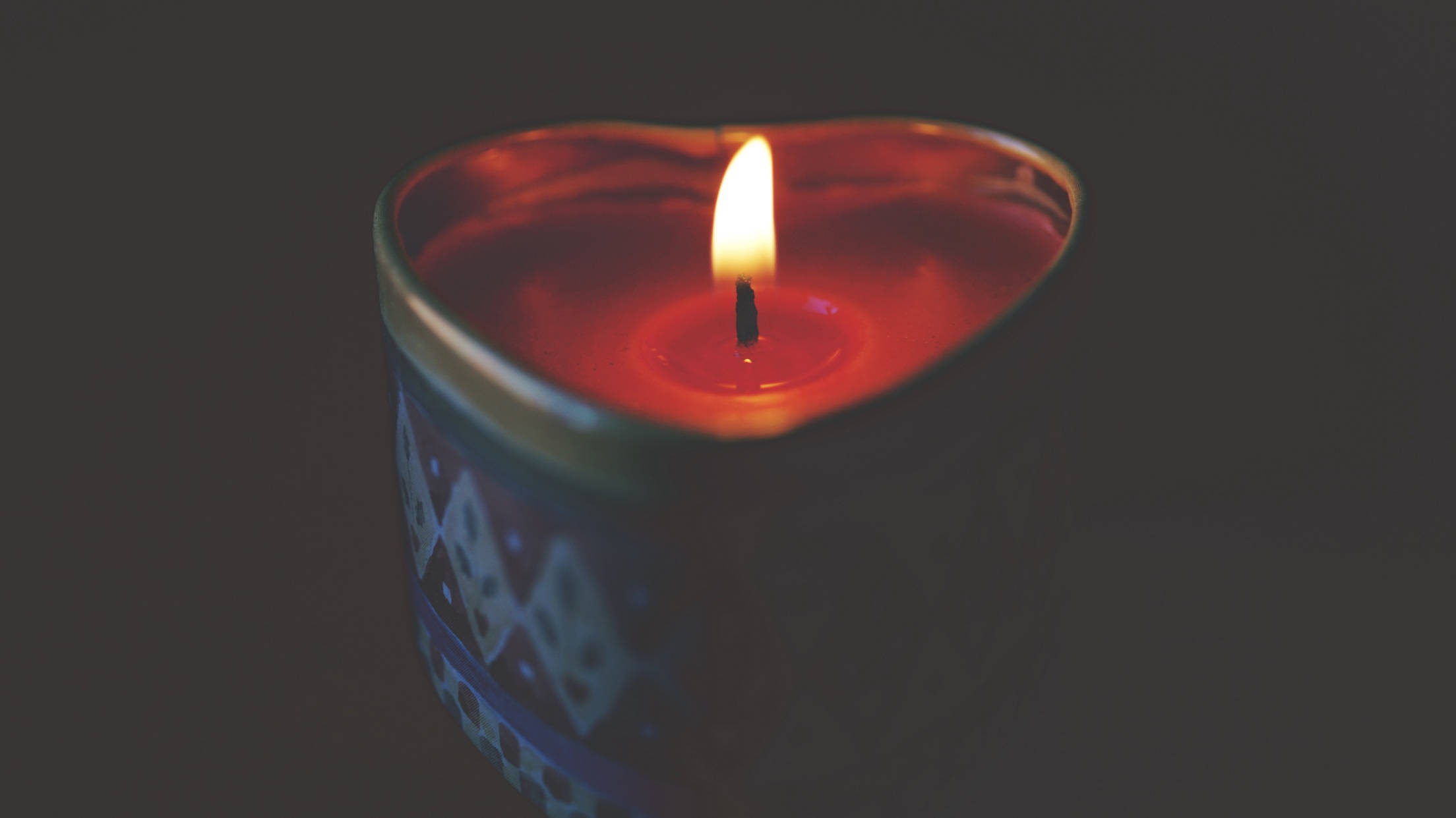蠟燭 桌布 壁紙 — 一顆心形蠟燭，火焰閃爍，在黑暗背景下散發出溫暖的紅光。