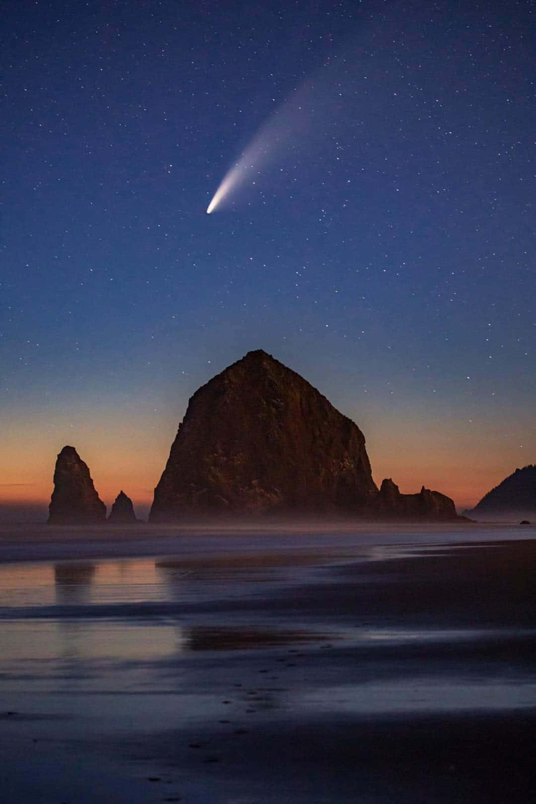 comet 桌布 壁紙 — 一顆明亮的彗星劃過滿天星斗的暮色天空，下方是霧氣籠罩的海岸岩石和寧靜如鏡的水面。