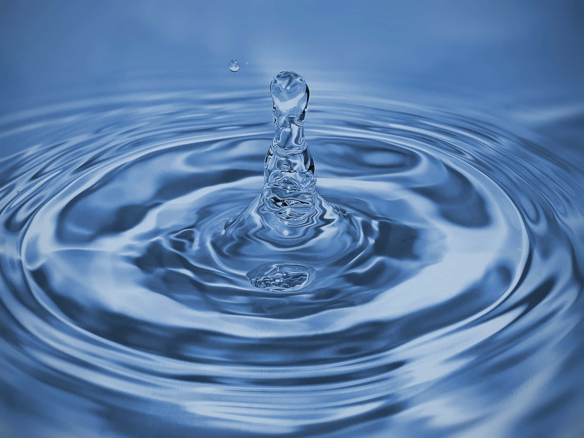 Water Drop 桌布 壁紙 — 一顆晶瑩剔透的水滴懸浮在半空中，在漣漪的藍色水面上形成同心圓波紋。
