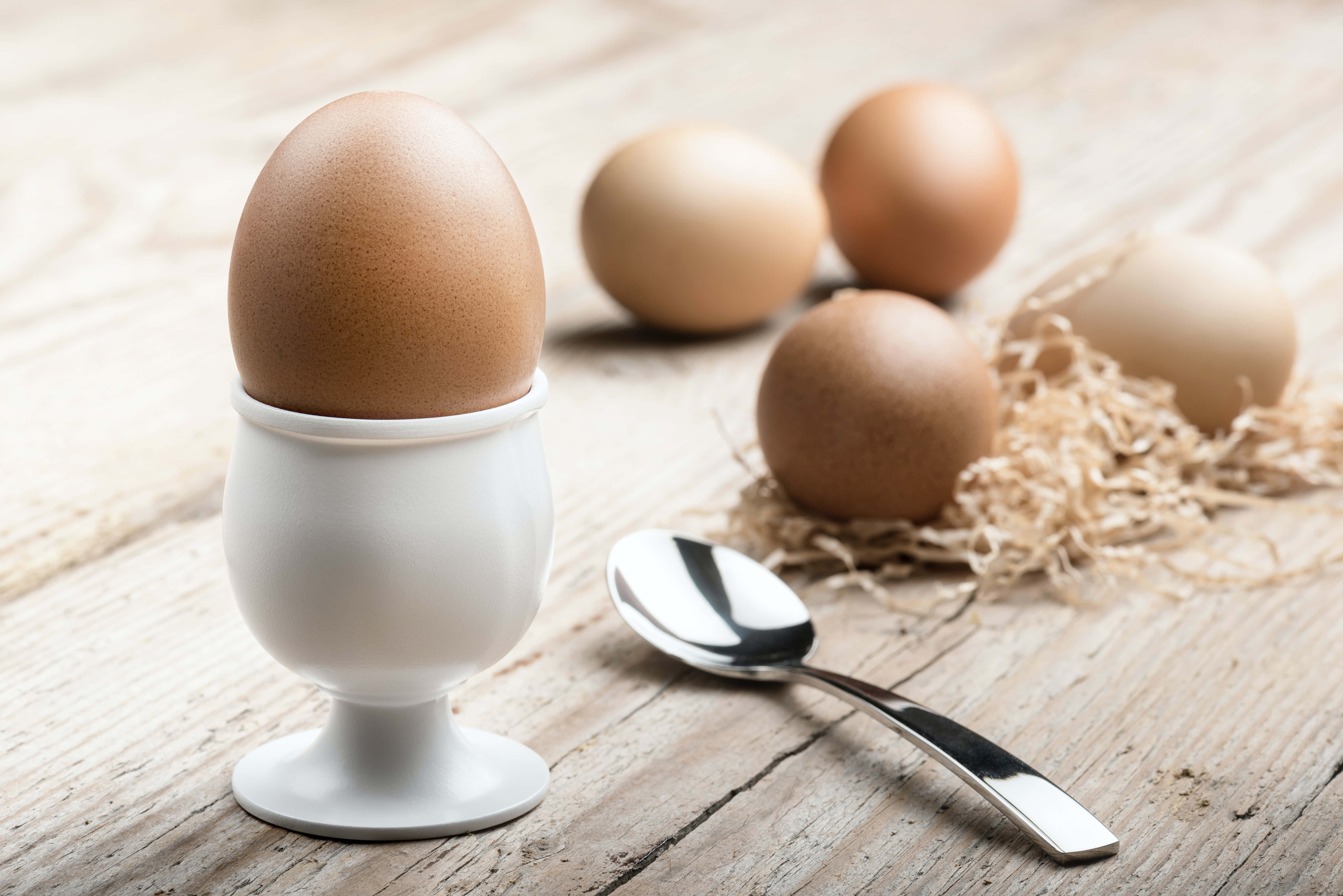 Brown Eggs 桌布 壁紙 — 一顆棕色雞蛋放在白色陶瓷蛋杯中，置於鄉村風木桌上，附近有散落的雞蛋和一把湯匙。