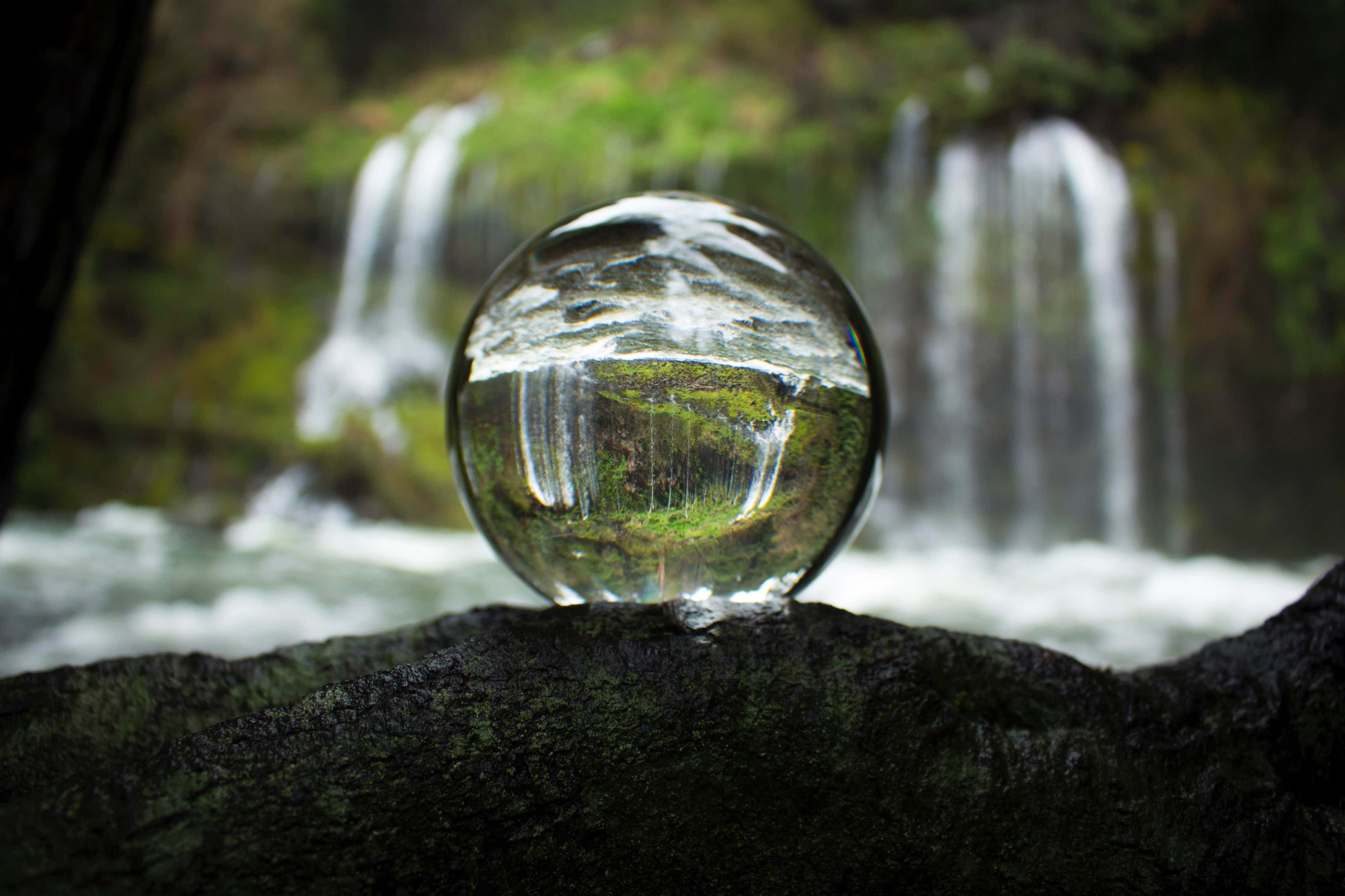 Crystal Ball 桌布 壁紙 — 一顆水晶球放在長滿苔蘚的岩石上，映照出模糊的瀑布和茂密的綠意，呈現寧靜的森林場景。