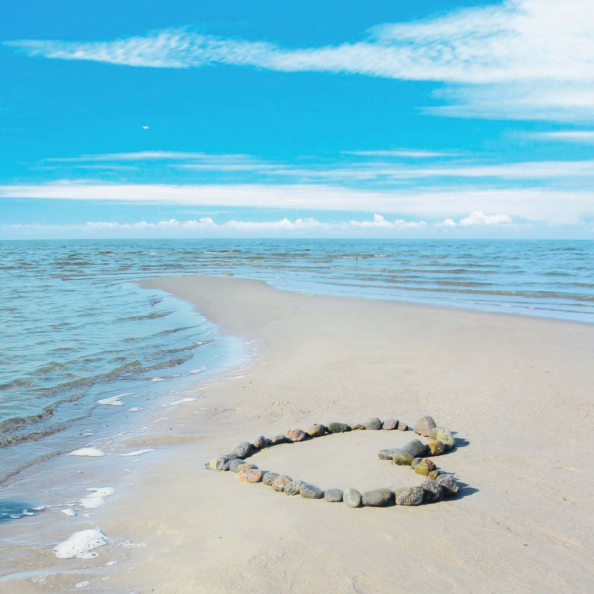 Beach Love 桌布 壁紙 — 一顆由光滑石頭組成的心形躺在沙灘上，溫柔的海浪在明亮的藍天下與地平線交匯。
