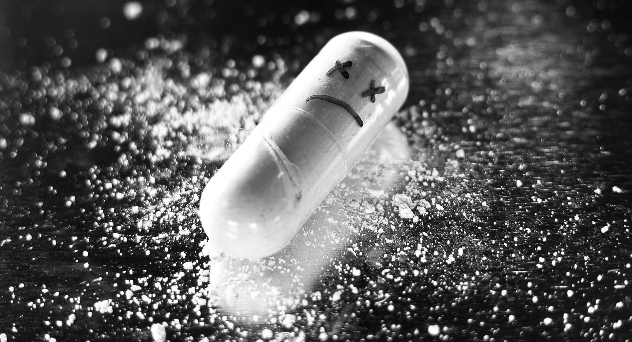 Monochrome 桌布 壁紙 — 一顆表情悲傷的藥丸躺在黑暗的表面上，周圍散落著白色粉末，呈現出鮮明的黑白照片效果。