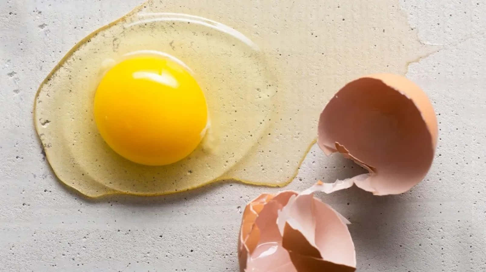 Egg 桌布 壁紙 — 一顆裂開的雞蛋，帶有鮮黃色的蛋黃和清澈的蛋白，在帶有斑點的灰色檯面上散開，旁邊有破碎的蛋殼半片。