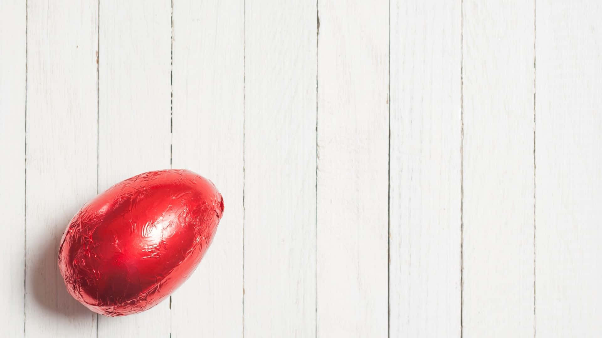 Easter Egg 桌布 壁紙 — 一顆閃亮的紅色箔紙包裹的巧克力蛋放在白色木板上。