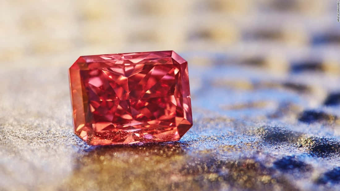 Red Diamond 桌布 壁紙 — 一顆閃耀的紅色長方形切割鑽石，在模糊的金色和藍色紋理表面上閃爍著火彩般的刻面。
