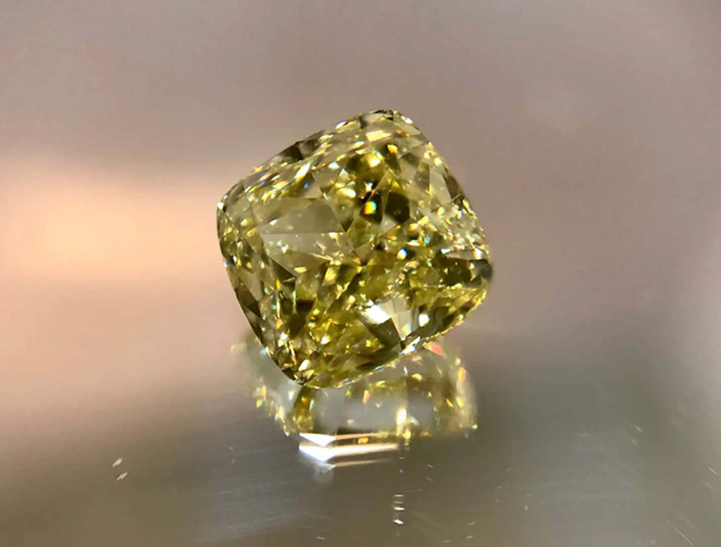 Yellow Diamond 桌布 壁紙 — 一顆閃耀的黃色枕形切割鑽石，在柔和的工作室燈光下，於反光金屬表面上閃爍著火焰般的刻面。