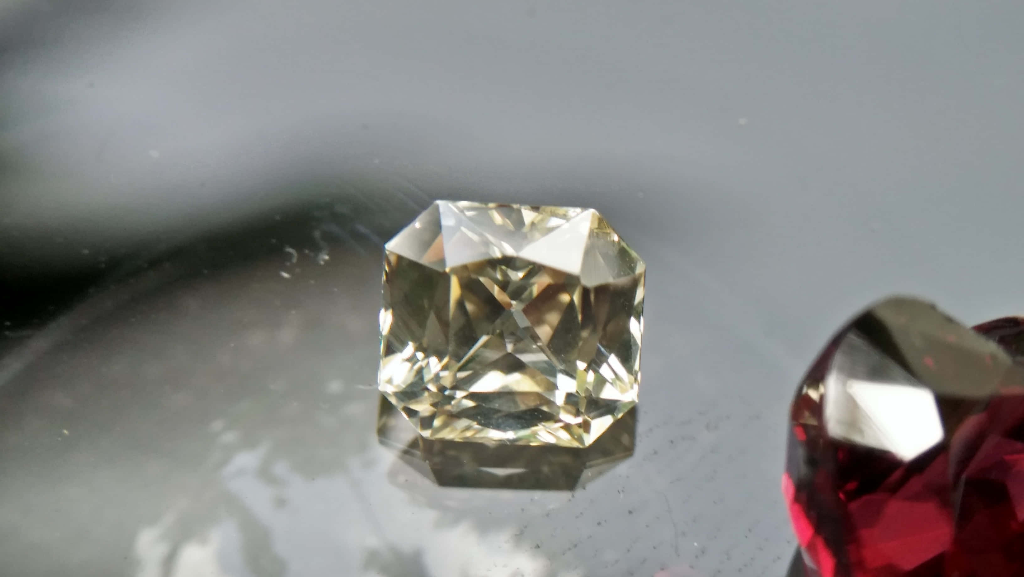 Yellow Sapphire 桌布 壁紙 — 一顆閃耀的黃色鑽石，帶有精確切割面，在反光的灰色表面上與一顆深紅色寶石相鄰閃爍。