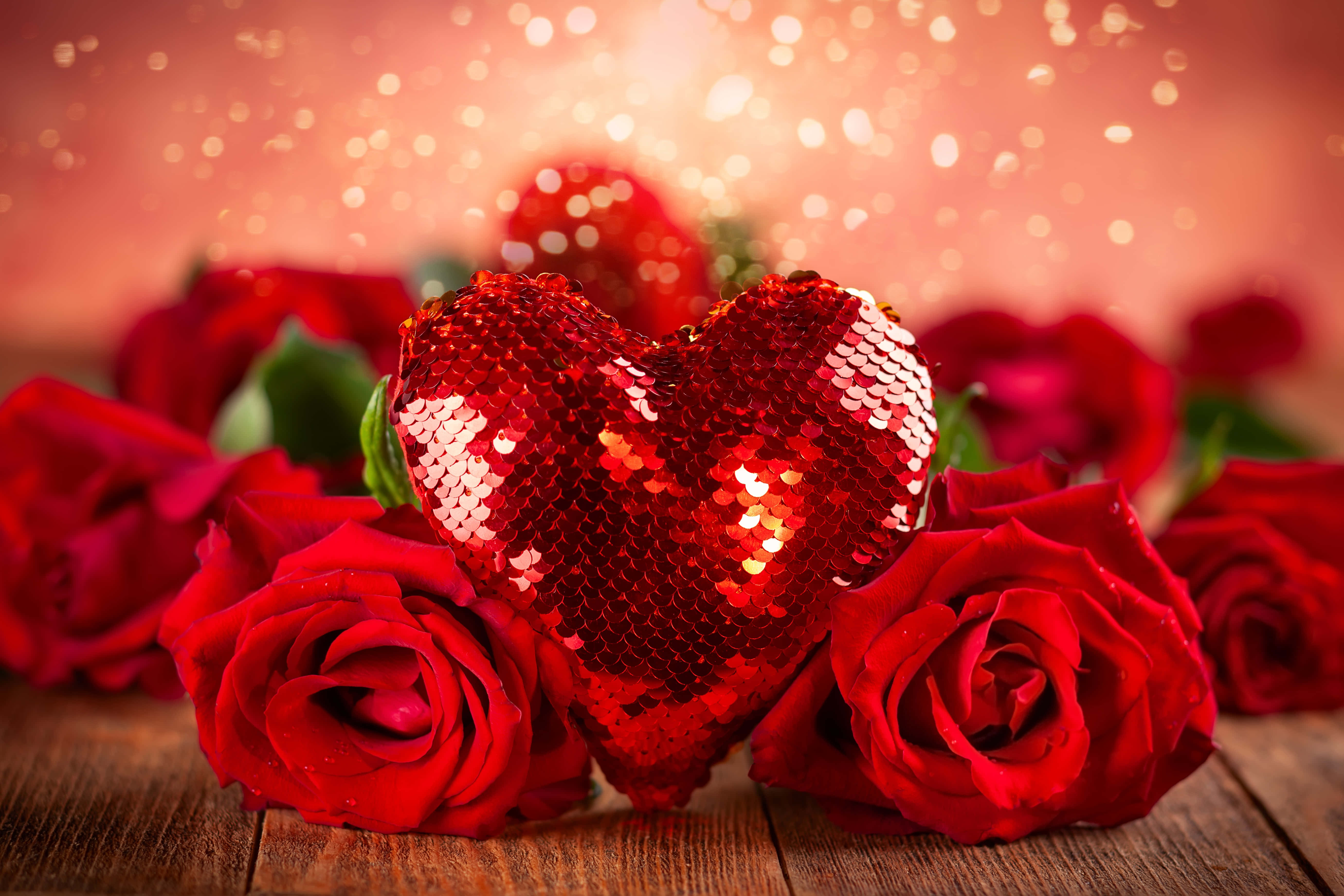 Valentines 桌布 壁紙 — 一顆閃閃發光的紅色亮片心形躺在鮮豔的紅玫瑰之間，置於質樸的木質表面上，伴隨著溫暖的散景燈光。
