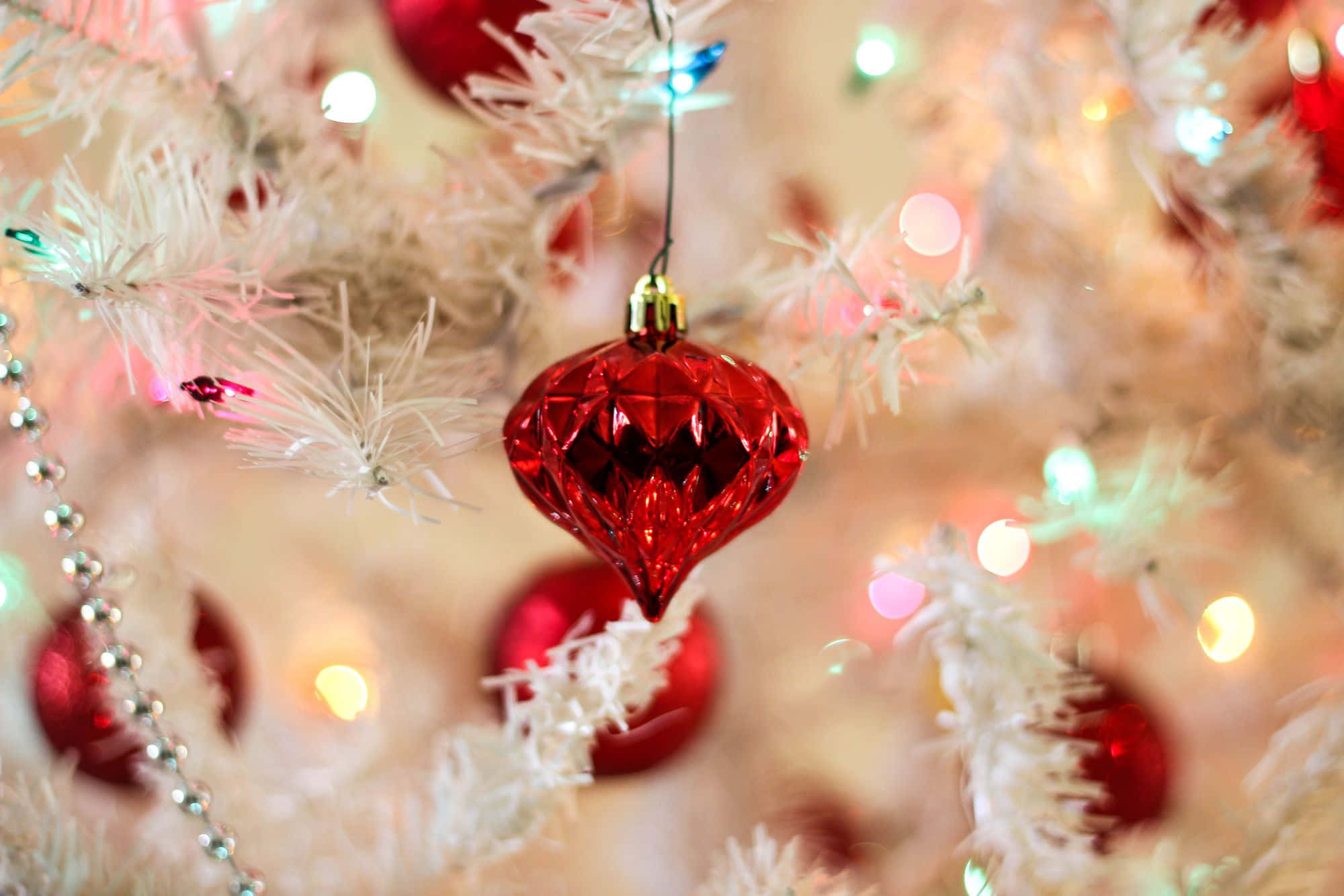 Teams Christmas 桌布 壁紙 — 一顆閃閃發光的紅色多面裝飾品掛在白色植絨聖誕樹上，樹上點綴著閃爍的彩色燈光。