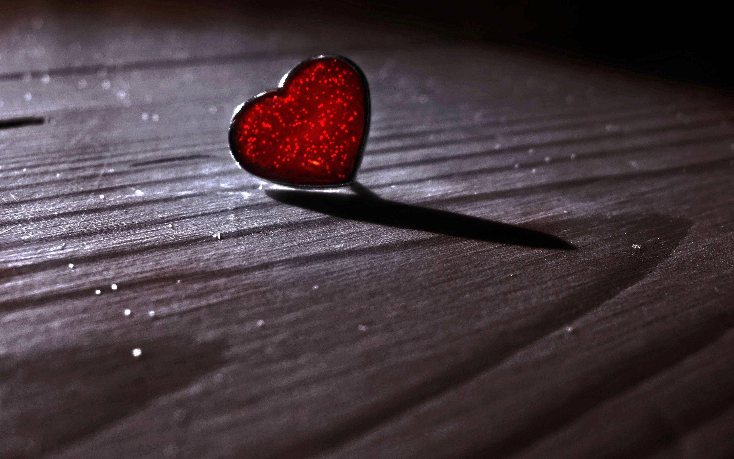 Valentines 桌布 壁紙 — 一顆閃閃發光的紅色心形別針獨自躺在深色木質表面上，在戲劇性的聚光燈下。