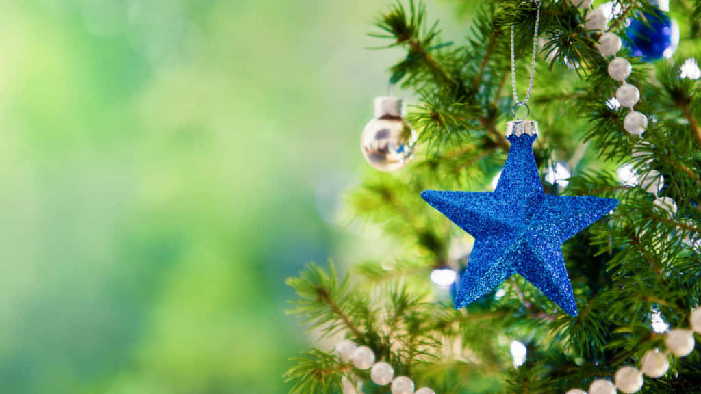 Christmas Star 桌布 壁紙 — 一顆閃閃發光的藍色星星裝飾品掛在茂盛的綠色松樹枝上，枝上點綴著銀色珠子和反光飾球。