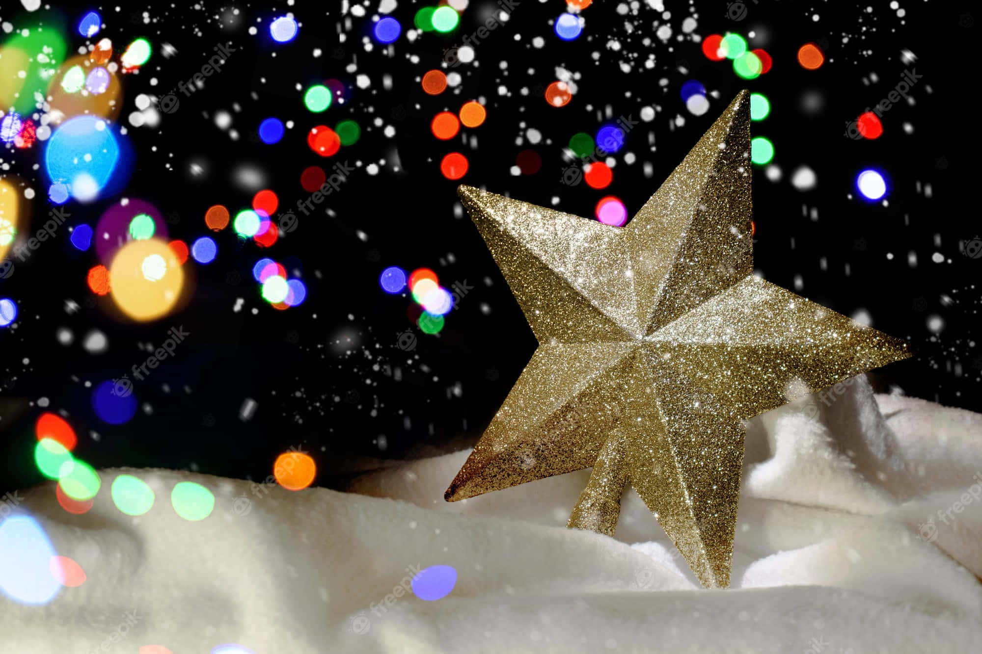 Christmas Star 桌布 壁紙 — 一顆閃閃發光的金色星星躺在蓬鬆的白雪上，背景是色彩繽紛的散景燈光和飄落的雪花。