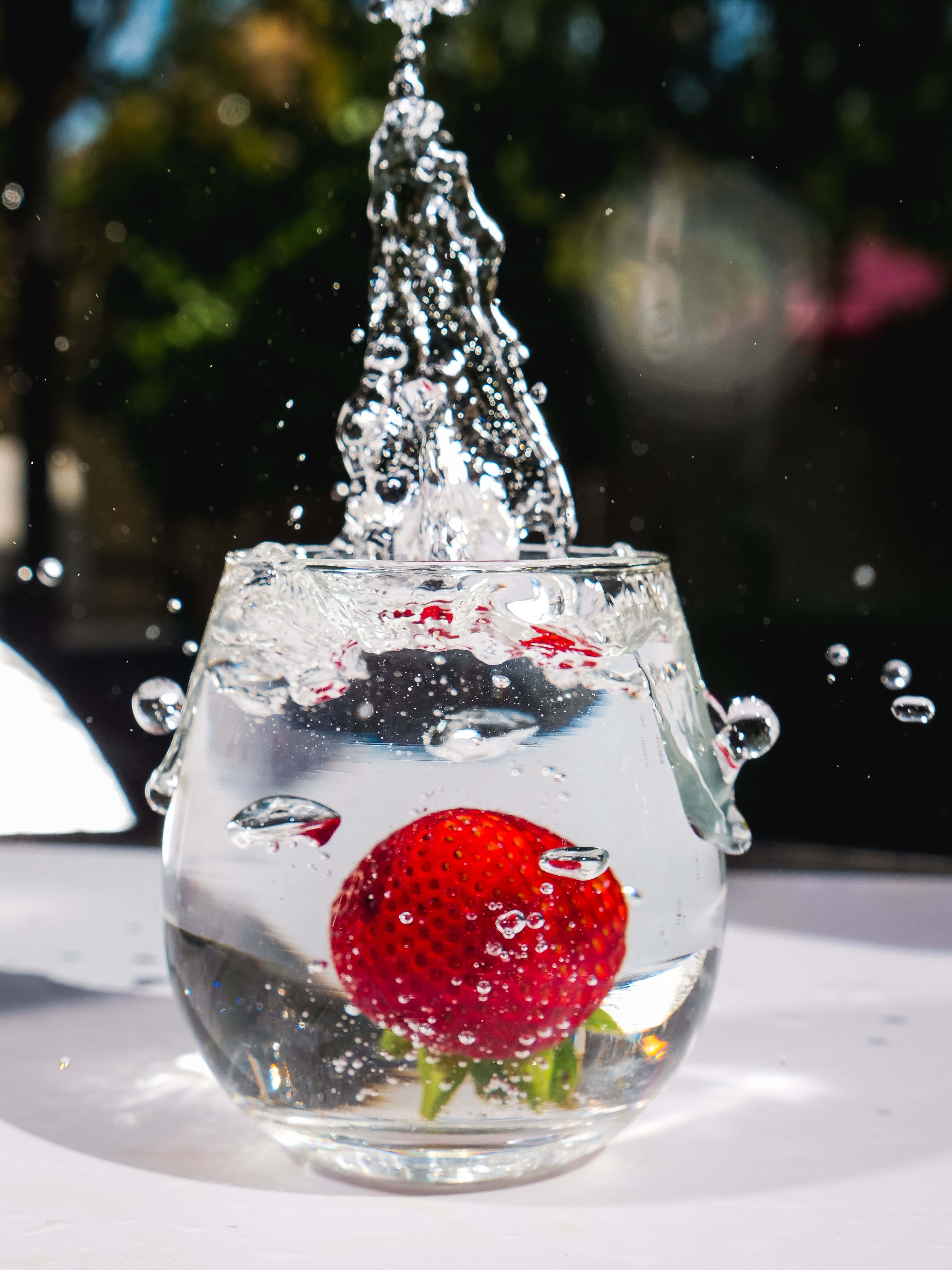 Glass Of Water 桌布 壁紙 — 一顆鮮紅的草莓掉進一杯水中，在模糊的綠色背景下，水滴在空中定格，形成動態的水花。