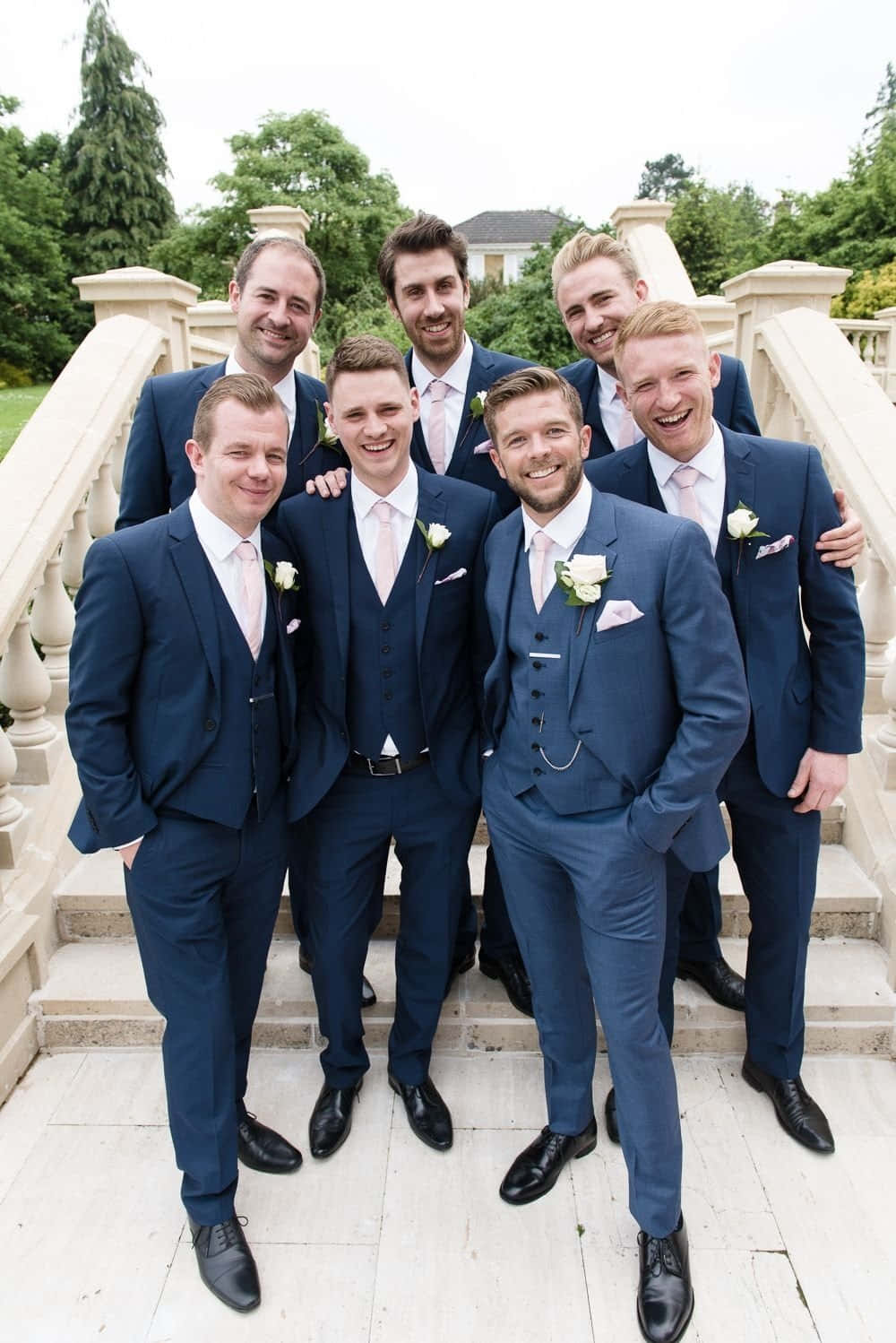 Groomsmen 桌布 壁紙 — 七位伴郎穿著相配的海軍藍西裝，佩戴白色襟花，在綠樹環繞的石階梯上一起大笑。