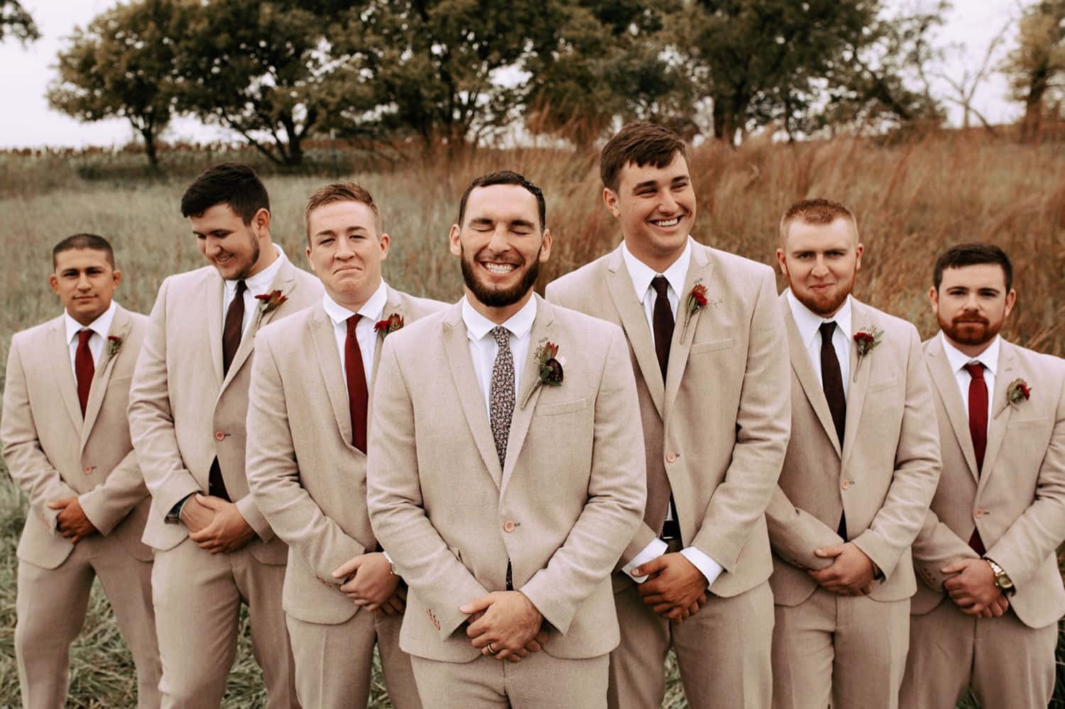 Groomsman 桌布 壁紙 — 七位伴郎身穿米色西裝和紅色領帶，在陰天的高草叢中微笑站立。
