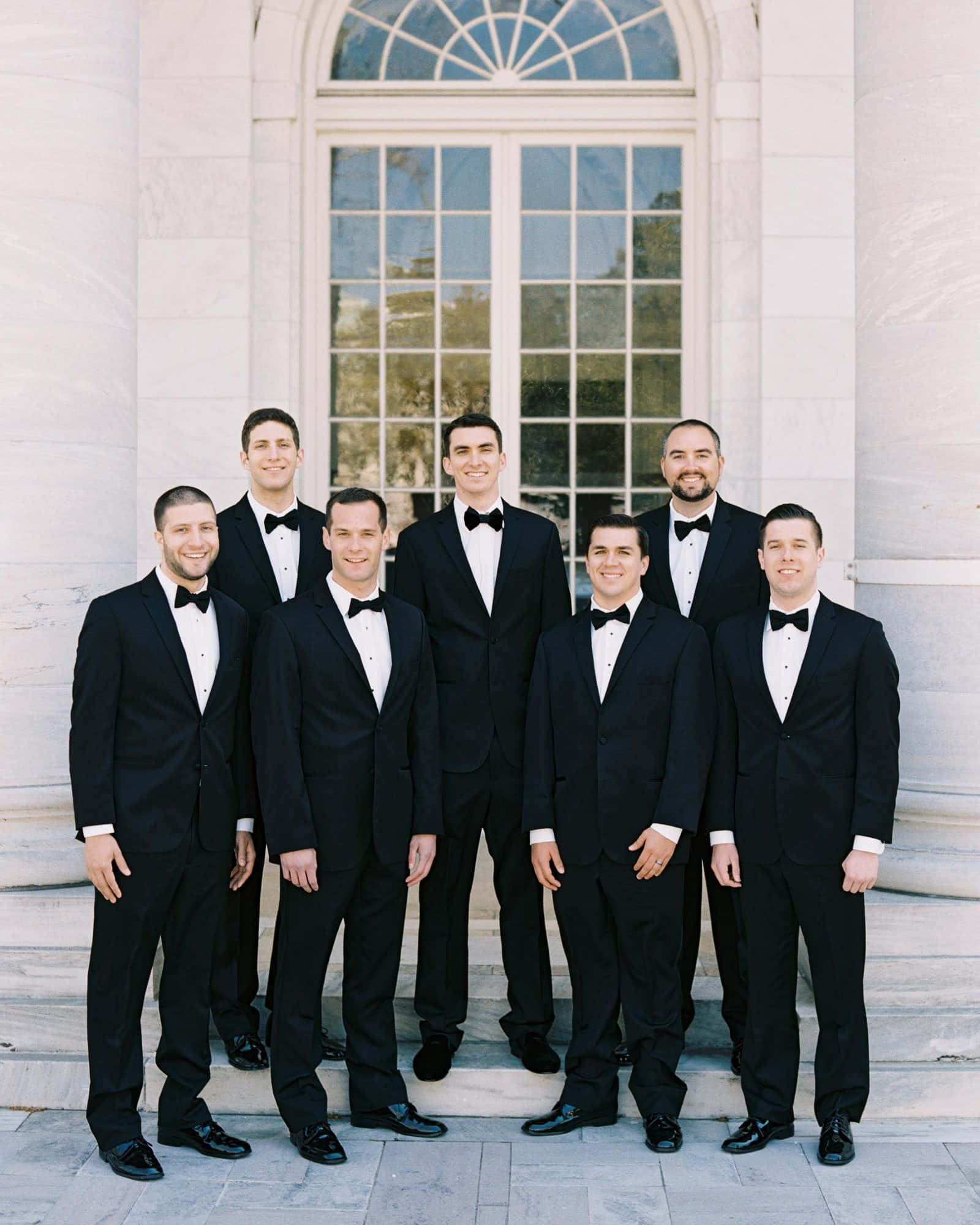 Groomsmen 桌布 壁紙 — 七位伴郎身穿黑色燕尾服配蝴蝶結，在白色柱式建築前的大理石階梯上正式擺姿勢，建築物有一個大型拱形窗戶。