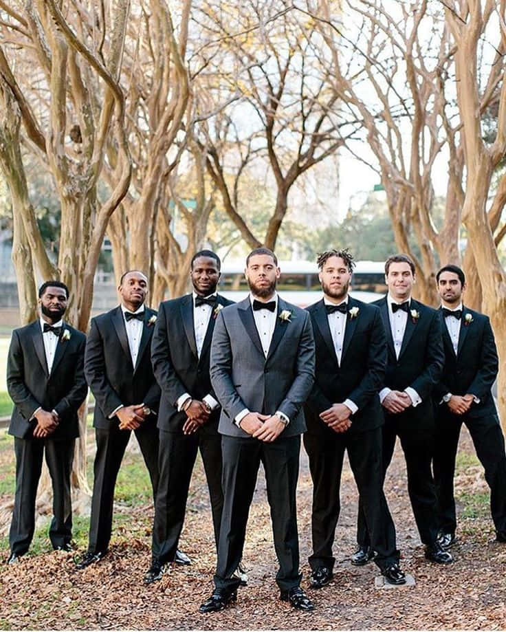 Groomsmen 桌布 壁紙 — 七位伴郎身穿黑色禮服配蝴蝶結，在光禿禿的樹下排成一排，地上有落葉。