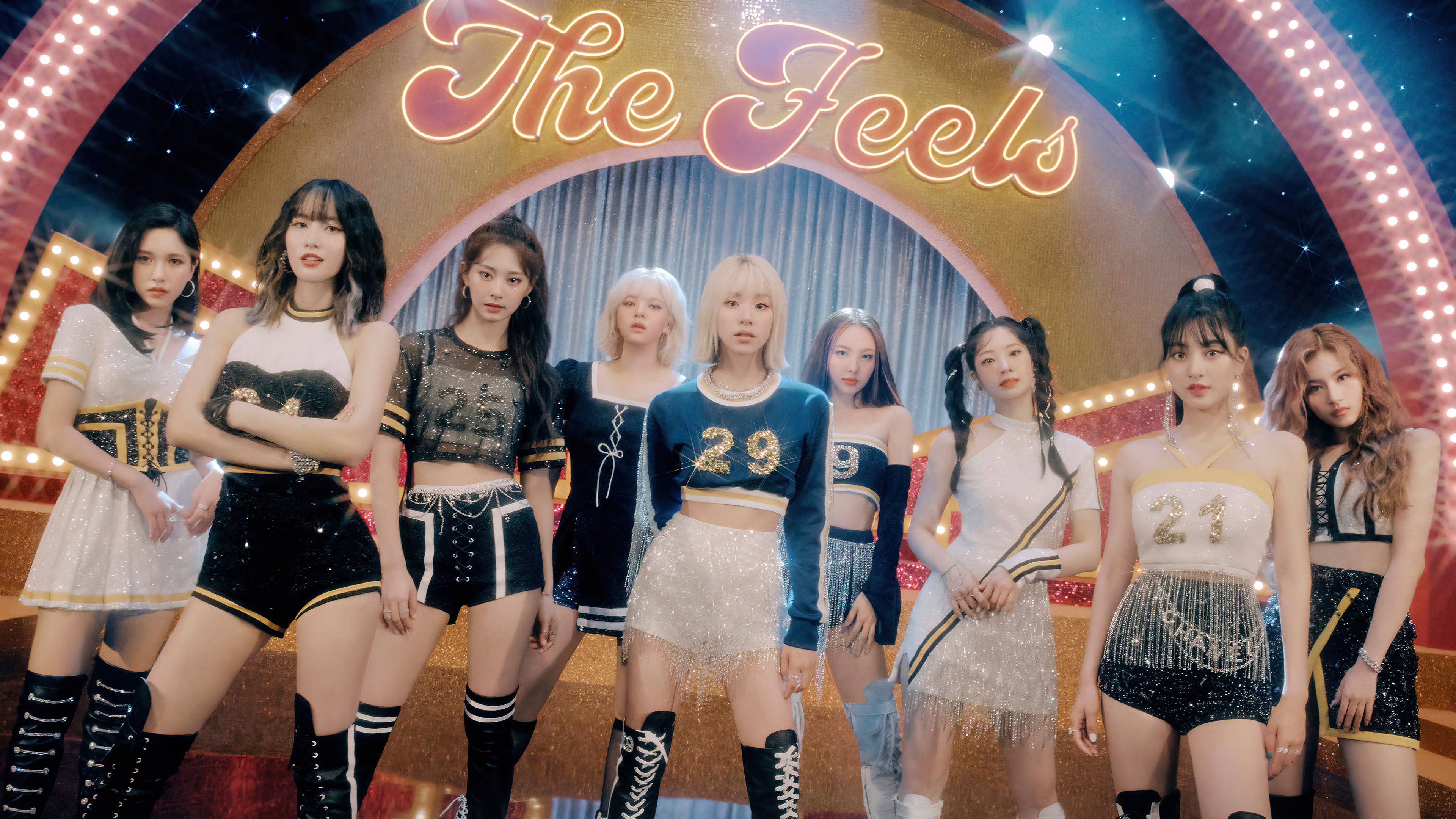 Kpop Aesthetic 桌布 壁紙 — 七位女性偶像在閃爍的啦啦隊服裝下，於發光的「The Feels」標誌前自信地擺姿勢，背景是充滿活力的舞台場景。