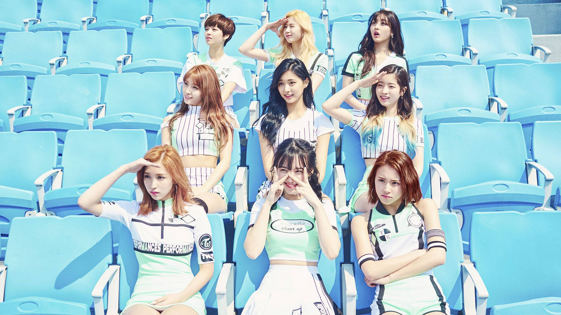 twice 桌布 壁紙 — 七位女性偶像穿著相配的運動服裝，在晴朗的日光下，在明亮的藍色體育場座位上俏皮地擺姿勢。