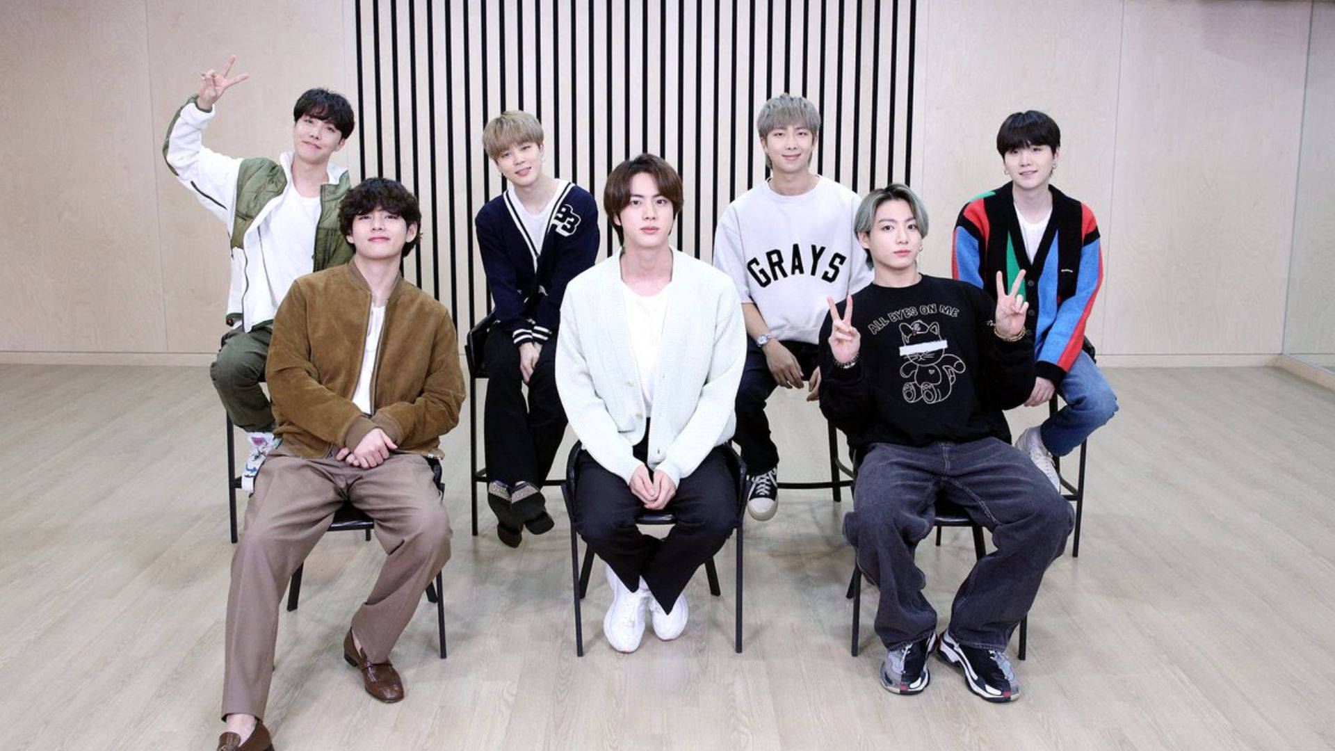 BTS 桌布 桌布 壁紙 — 七位年輕男士穿著休閒服裝，在工作室的條紋牆前坐在椅子上愉快地擺姿勢。
