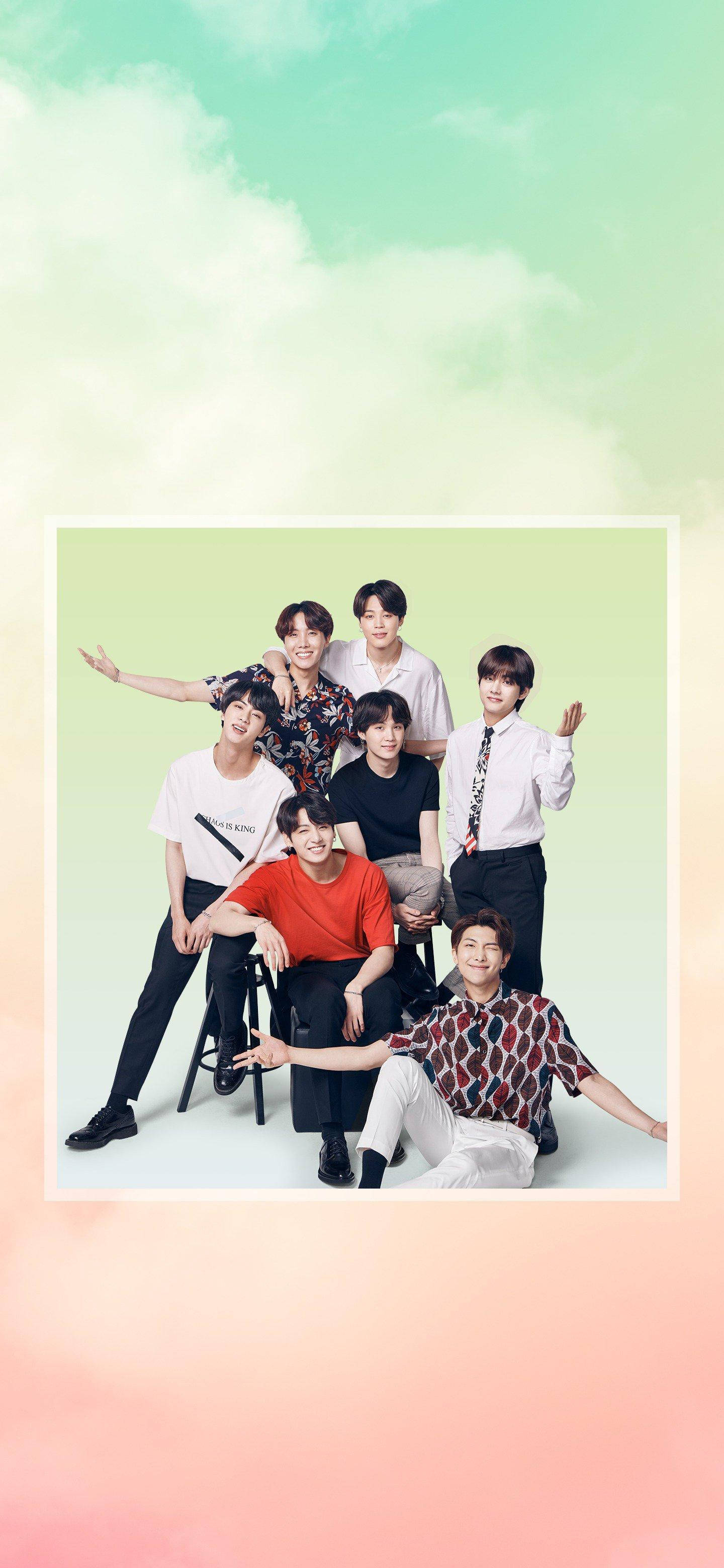 BTS 桌布 壁紙 — 七位年輕男士穿著時尚休閒服裝，在柔和的粉彩色漸變背景前，伴隨著蓬鬆的雲朵，開心地擺姿勢。