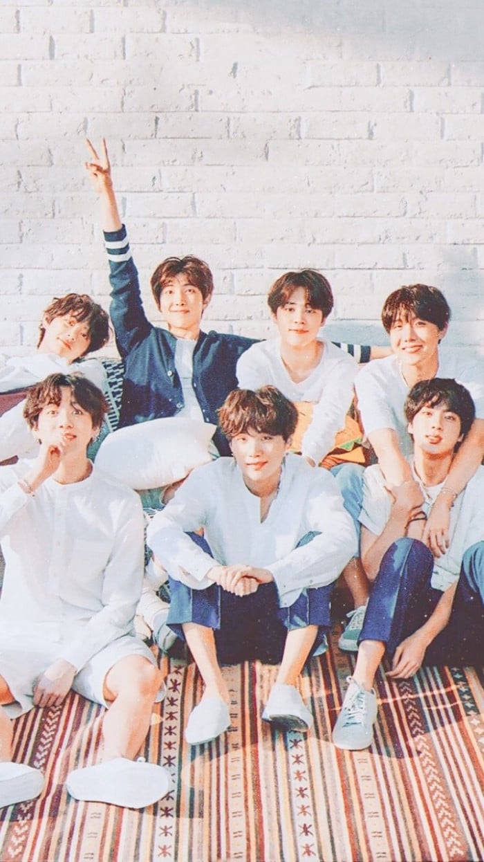 BTS 桌布 壁紙 — 七位年輕男士身穿休閒服裝，在白色磚牆前的圖案地毯上愉快地擺姿勢。
