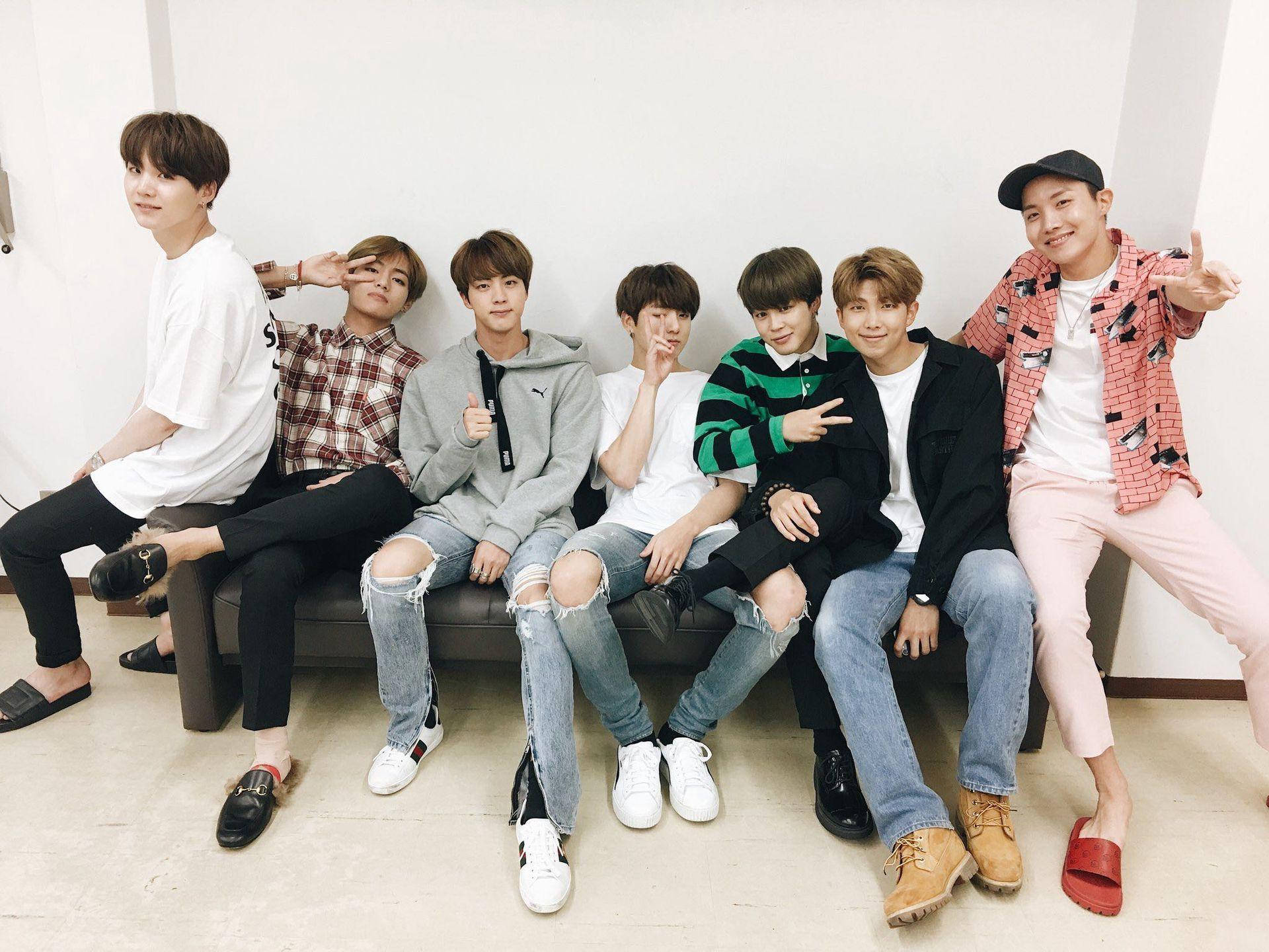 Cute Bts 桌布 壁紙 — 七位年輕男士身穿休閒服裝，在白色牆壁前的沙發上愉快地擺姿勢。
