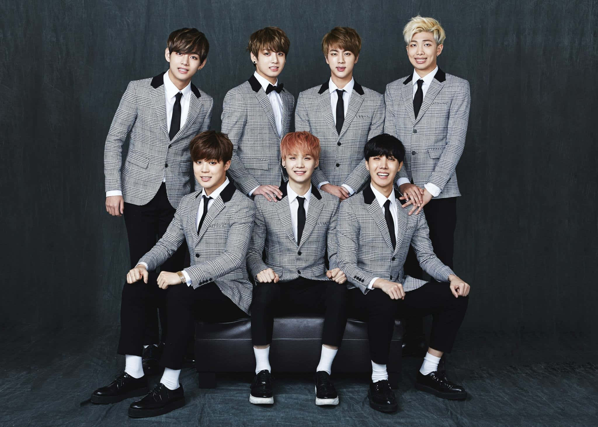 BTS 桌布 壁紙 — 七位年輕男士身穿相配的灰色格紋西裝，自信地坐在黑色長椅上，背景是深色紋理背景。