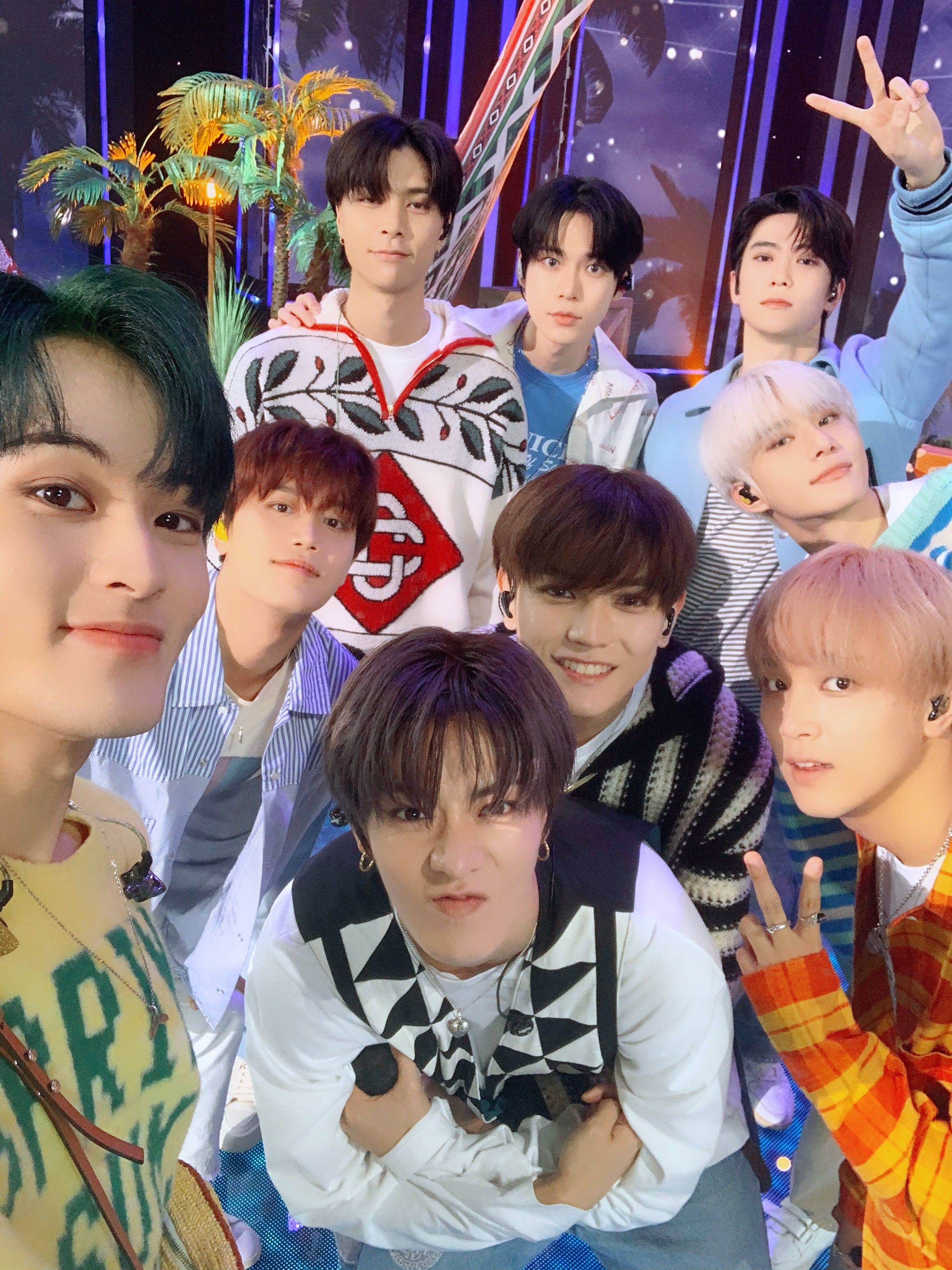 Nct 桌布 壁紙 — 七位年輕男子，頭髮色彩繽紛，穿著時尚，在充滿活力的舞台場景中，棕櫚樹和閃爍燈光下愉快地擺姿勢。