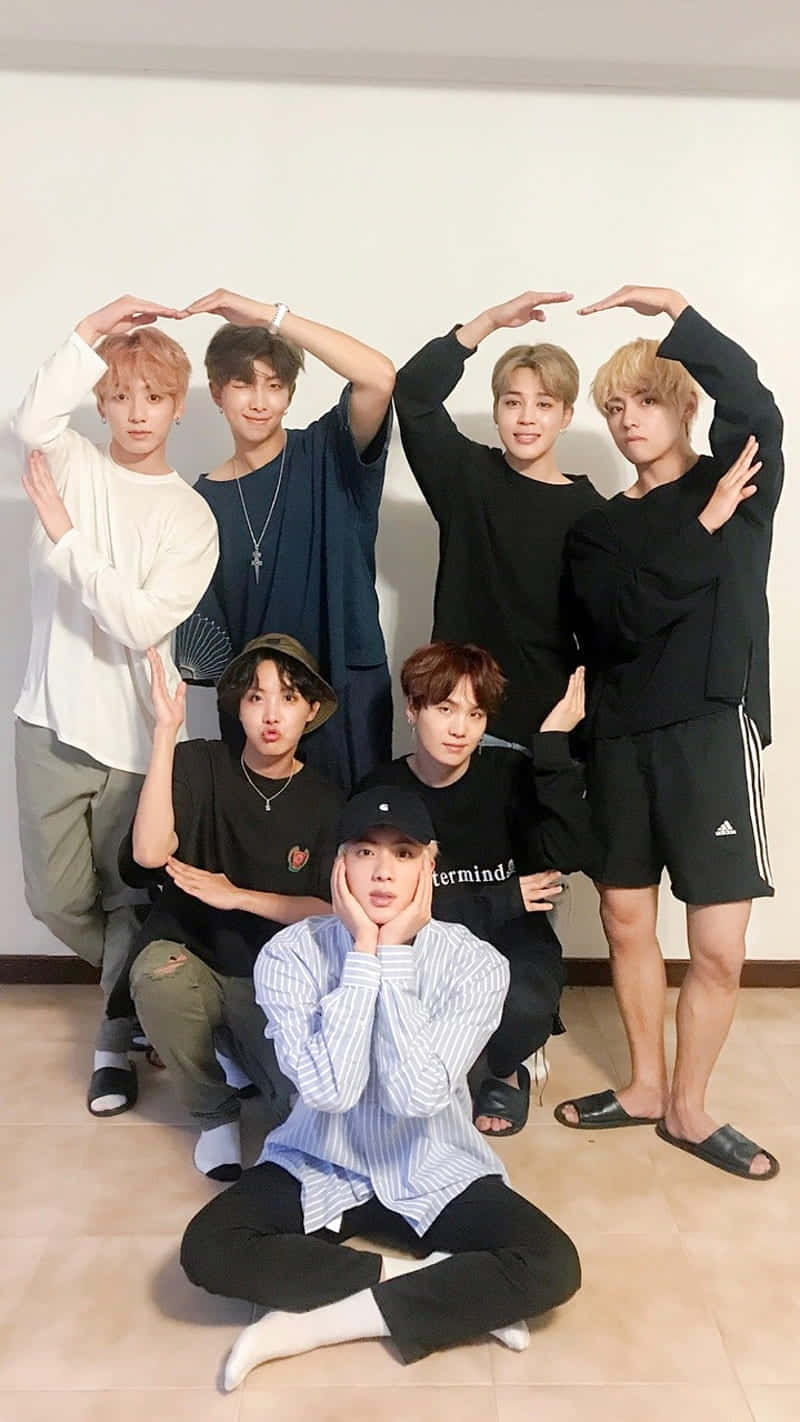 BTS 桌布 壁紙 — 七位年輕男子在室內對著素色牆壁，雙手在頭頂做出形狀，擺出俏皮的姿勢。