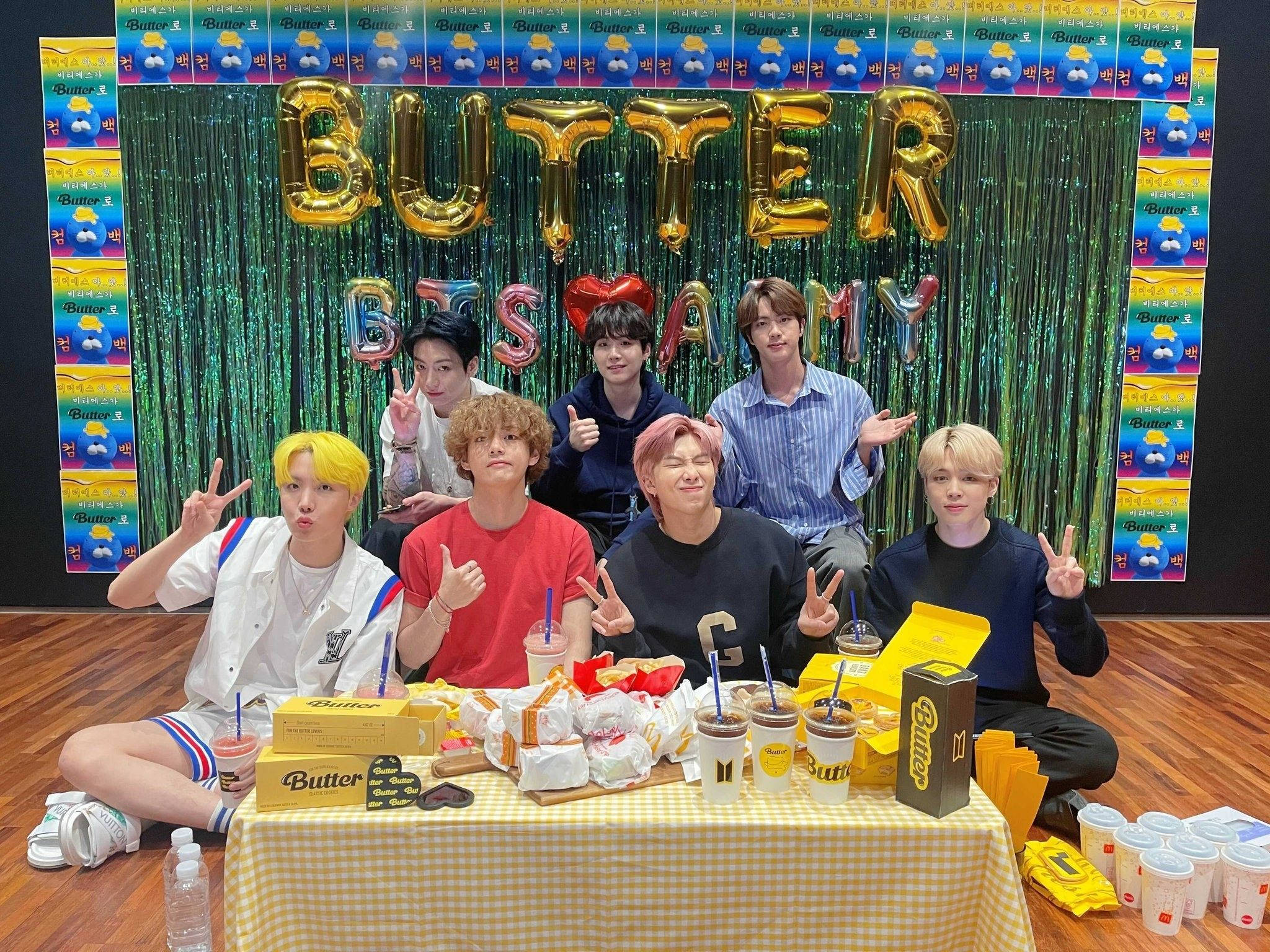 BTS 2021 桌布 壁紙 — 七位年輕男子在閃爍的綠色背景下，愉快地擺姿勢，身後是一張擺滿奶油主題小食和飲品的桌子。